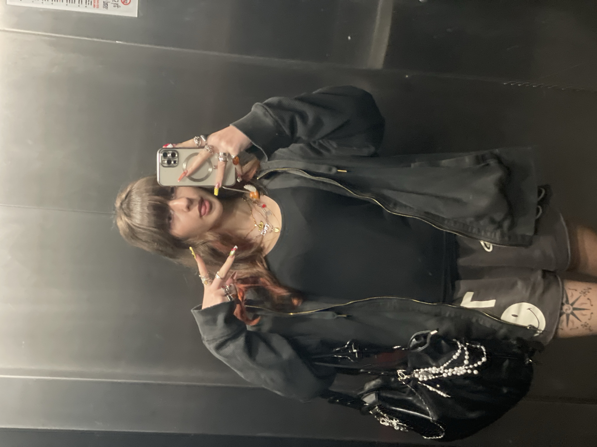 A young woman is taking a selfie in what appears to be an elevator. She is wearing a black top, a dark jacket, and grey shorts with a white smiley face design. She has multiple rings on her fingers and a necklace. She is holding a smartphone in her right hand to take the picture, making a peace sign with her left hand. A black purse with pearl embellishments is slung over her shoulder. A compass rose tattoo is visible on her leg. The elevator's interior is plain, with a metallic finish. The image lacks enough information to determine the exact location in the city of Raanana, Israel.