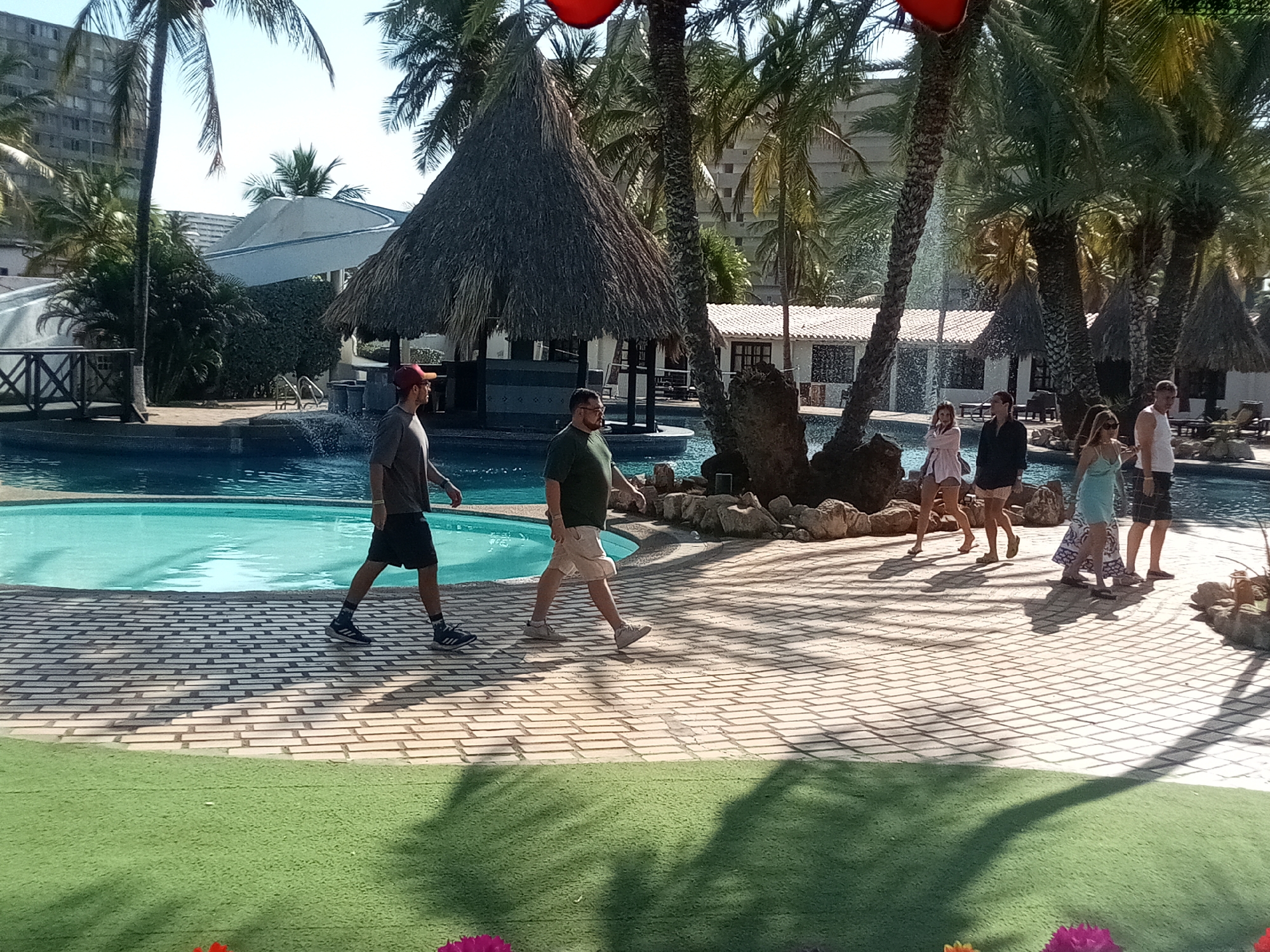This image shows a scene in what appears to be a resort in Porlamar, Venezuela. A swimming pool is central to the scene, bordered by brick walkways, artificial green turf, palm trees, and thatched-roof structures. Two men are walking along the left side of the pool: one is wearing a red baseball cap, gray shirt, and black shorts, and the other is wearing a green shirt and khaki shorts. On the other side of the pool, four people are walking in the opposite direction: a woman in a light pink outfit, a woman in a black outfit, a woman in a light blue dress, and a man in a white tank top and dark shorts. There is a water fountain feature by the edge of the pool near them. The background includes resort buildings and tall palm trees. The day is sunny, casting shadows from the palm trees across the brick walkway.
