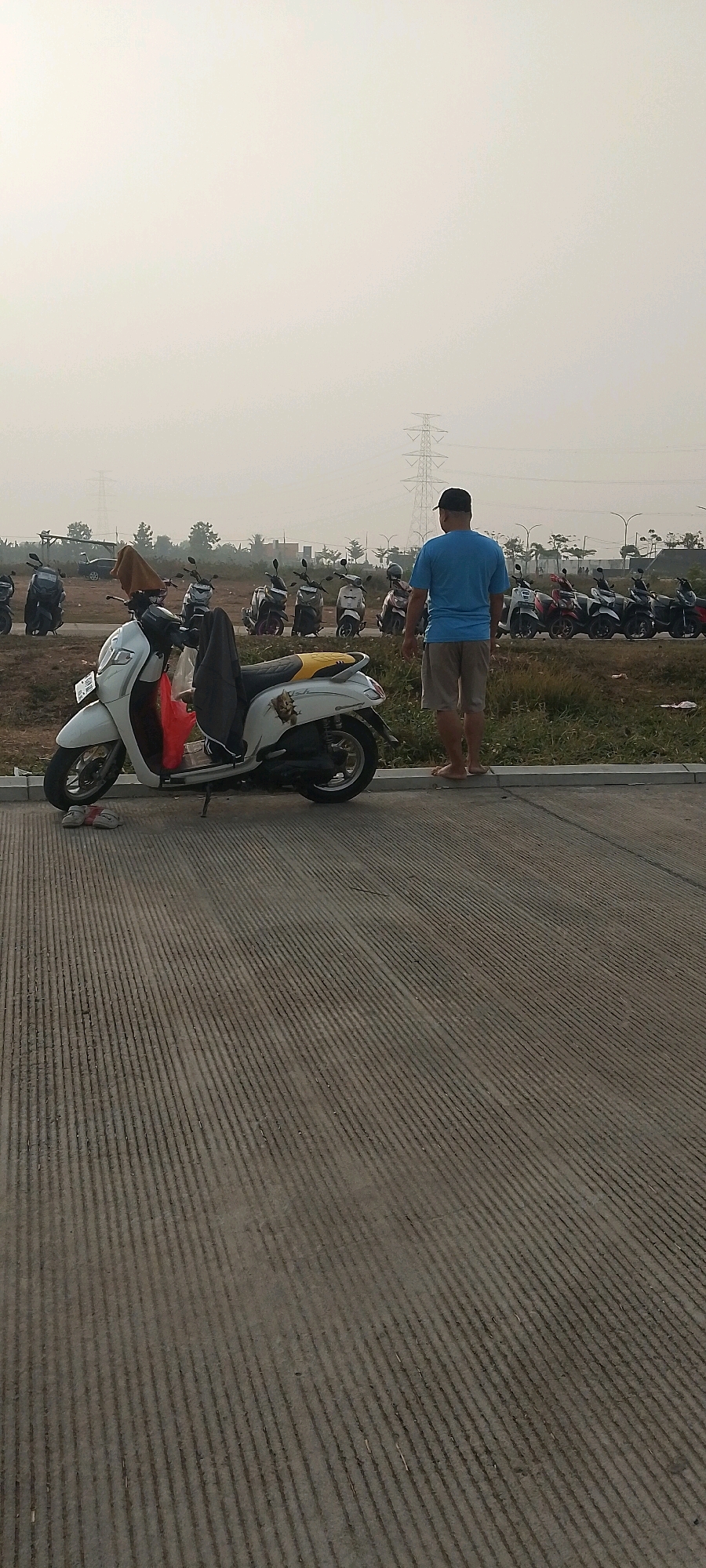 A medium shot shows a man wearing a blue t-shirt and beige shorts standing barefoot on a raised concrete curb. He faces away from the camera, looking towards a line of parked motorbikes. In the foreground, a white scooter is parked on the concrete surface. Behind the man, a row of various motorbikes are parked in a grassy area. In the background, a large electricity pylon is visible against a hazy sky. The overall environment appears to be an outdoor parking area.