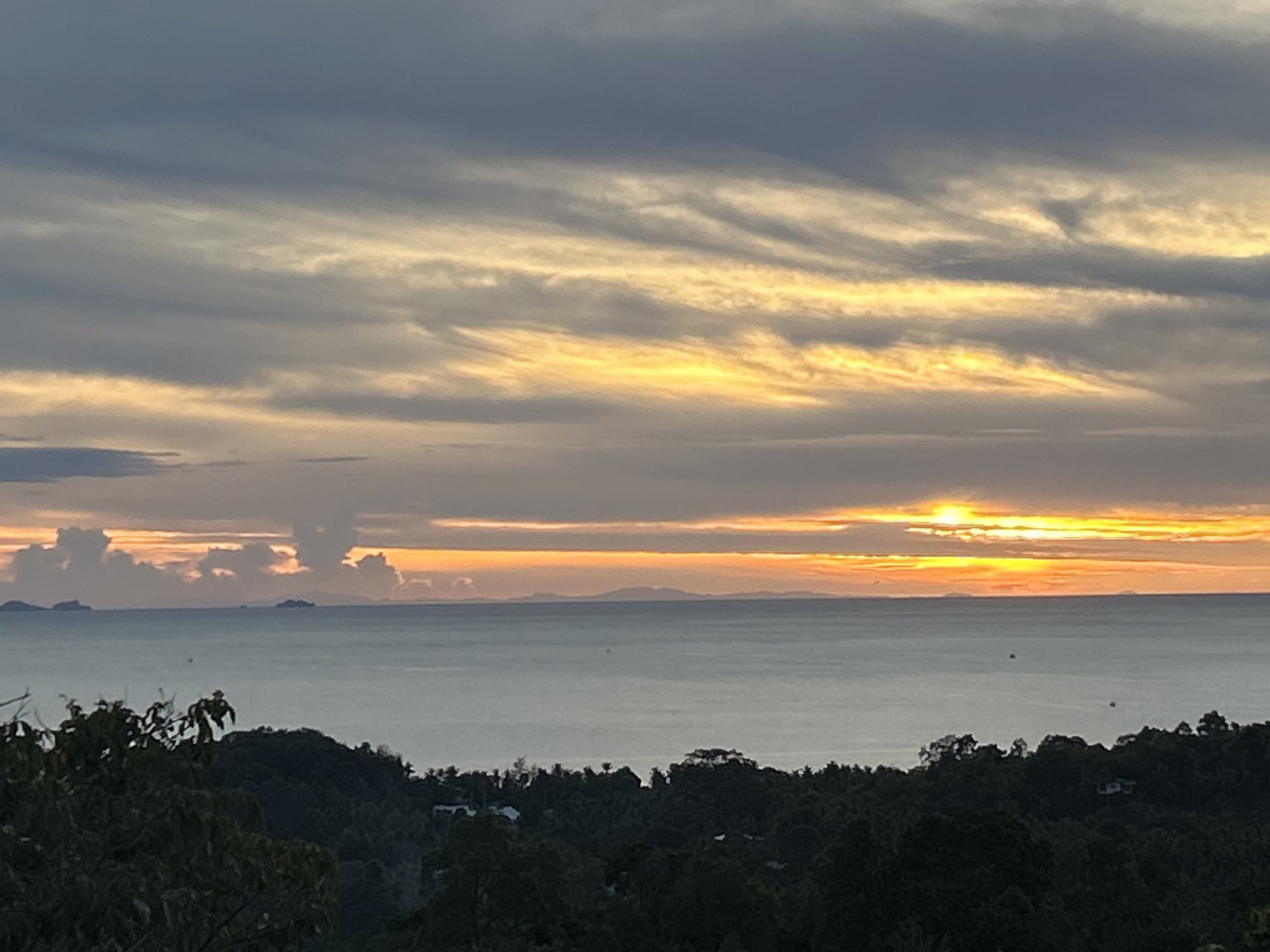 The image depicts a seascape at sunset in Baan Sri Thanu, Thailand. In the foreground, a lush, dark green forest occupies the lower portion of the frame. Above the forest, the sea stretches across the middle of the image, with distant islands faintly visible on the horizon. The sky dominates the upper half of the image, featuring a mix of dark, gray clouds and streaks of vibrant yellow and orange hues emanating from the setting sun. The sun's light reflects on the water's surface.