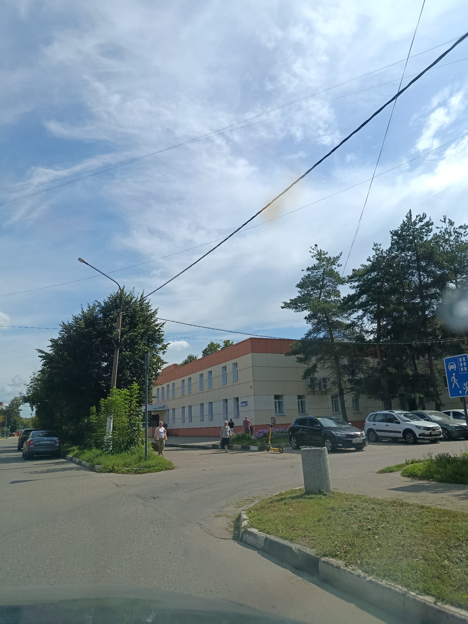 The image captures a street scene in Orekhovo-Zuyevo, Russia, on a clear, sunny day. In the background, a two-story building with a cream-colored facade and an orange band beneath the roofline is visible. The building has several windows with white frames. To the left of the building, a large deciduous tree with dark green foliage stands prominently, obscuring part of the structure. Further to the right, several tall pine trees are interspersed with the building.
In the foreground, a paved road curves through the scene. Several cars are parked along the right side of the road, including a white SUV, a dark hatchback, and other vehicles further down. A blue and white traffic sign depicting a car and pedestrians is also visible near the pine trees.
On the sidewalk in front of the building, several people are present. A woman in a light-colored top and dark pants walks towards the left, carrying a bag. Another woman in a light-colored dress and carrying a bag is seen further to the right. A man in a red shirt is also visible. A yellow electric scooter is parked near the sidewalk.
The sky is blue with scattered white clouds, and power lines are strung across the upper portion of the image. The overall impression is of a typical urban street environment during daylight hours.