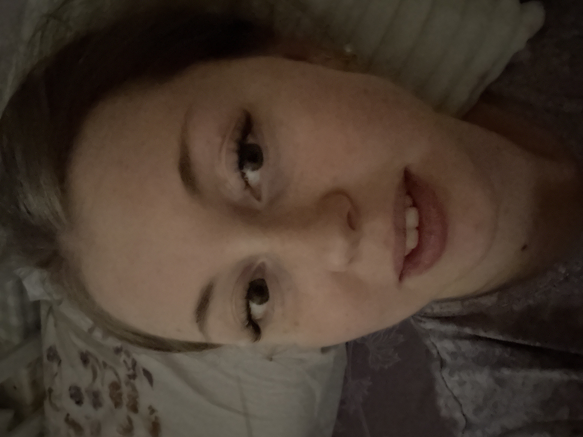 A person is lying down, possibly in a bed, in an unknown location. The person has fair skin, dark eyes, and brown hair. They appear to be looking at the camera. The background includes pillows and fabric.