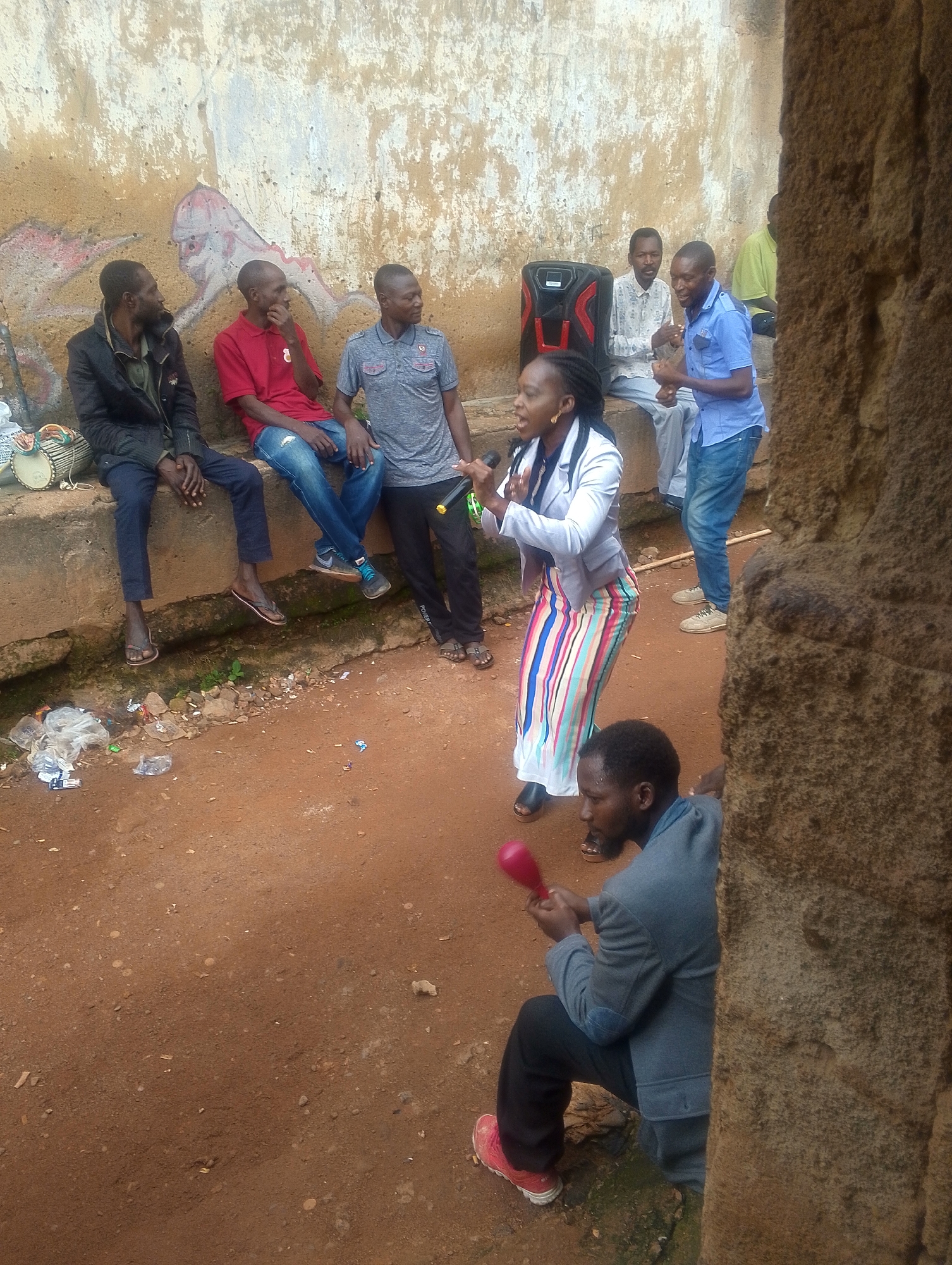 In an outdoor, earthy environment, a group of individuals are gathered near a textured, possibly ruined, concrete wall with some faded murals. A woman in a white blazer and a colorful striped skirt stands at the center, holding a microphone and a maraca, appearing to be performing or speaking. To her left, a black portable speaker sits on a concrete ledge. Several men are seated or standing around, observing. One man in a blue shirt and jeans is standing and gesturing, while others are seated on the ledge, including a man in a black jacket, a man in a red polo shirt, and a man in a gray t-shirt. Another man is partially visible in the foreground, crouched down and also holding a maraca. The ground is covered in reddish-brown soil with scattered debris.