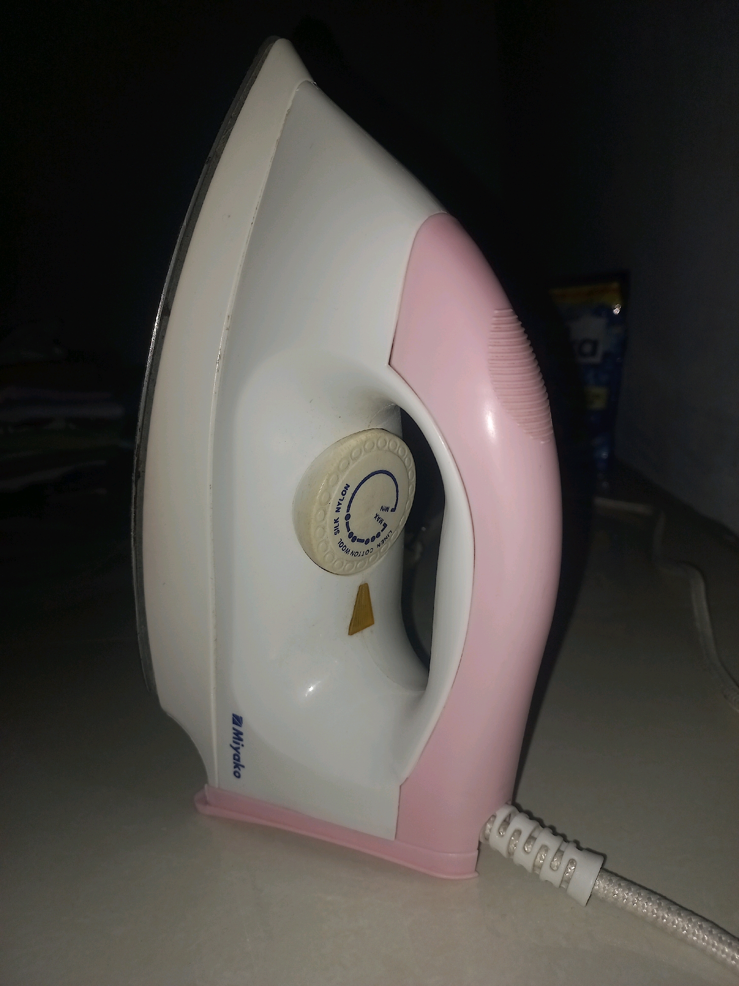 An electronic iron with a white and pink body sits on a light-colored surface in what appears to be a dimly lit indoor environment. The iron has a dial with fabric settings such as "SILK", "NYLON", "WOOL", "COTTON", and "LINEN" indicated, along with "MIN" and "MAX" settings. The brand name "Miyako" is visible in blue text on the side of the iron. The power cord, white and braided, is connected to the iron and extends out of frame. In the blurred background, a blue and yellow object is visible. The overall scene suggests a household setting.