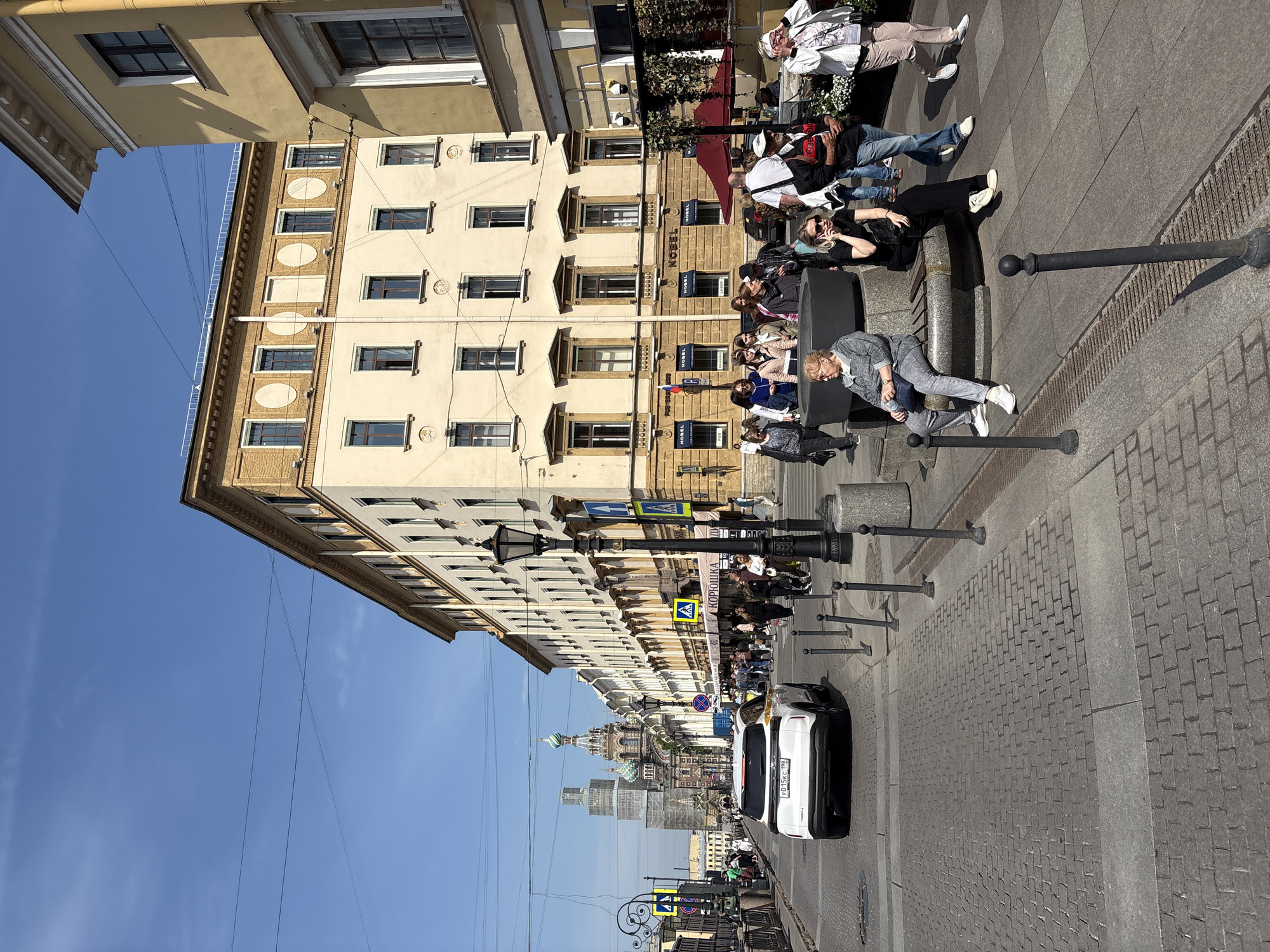 The image depicts a street scene in Saint Petersburg, Russia. The perspective is from the side of a street, looking down its length. On the right is a building with multiple windows and architectural details. A group of people are gathered along the sidewalk, some seated on a ledge, appearing relaxed or engaged in conversation. The building behind them has "Nobel" signs affixed to it. The foreground features metal poles spaced along the sidewalk edge. A white vehicle is parked on the street. Traffic and pedestrians are visible further down the street, and in the distance, the distinctive domes of the Church of the Savior on Spilled Blood are visible. The sky is a clear blue.
