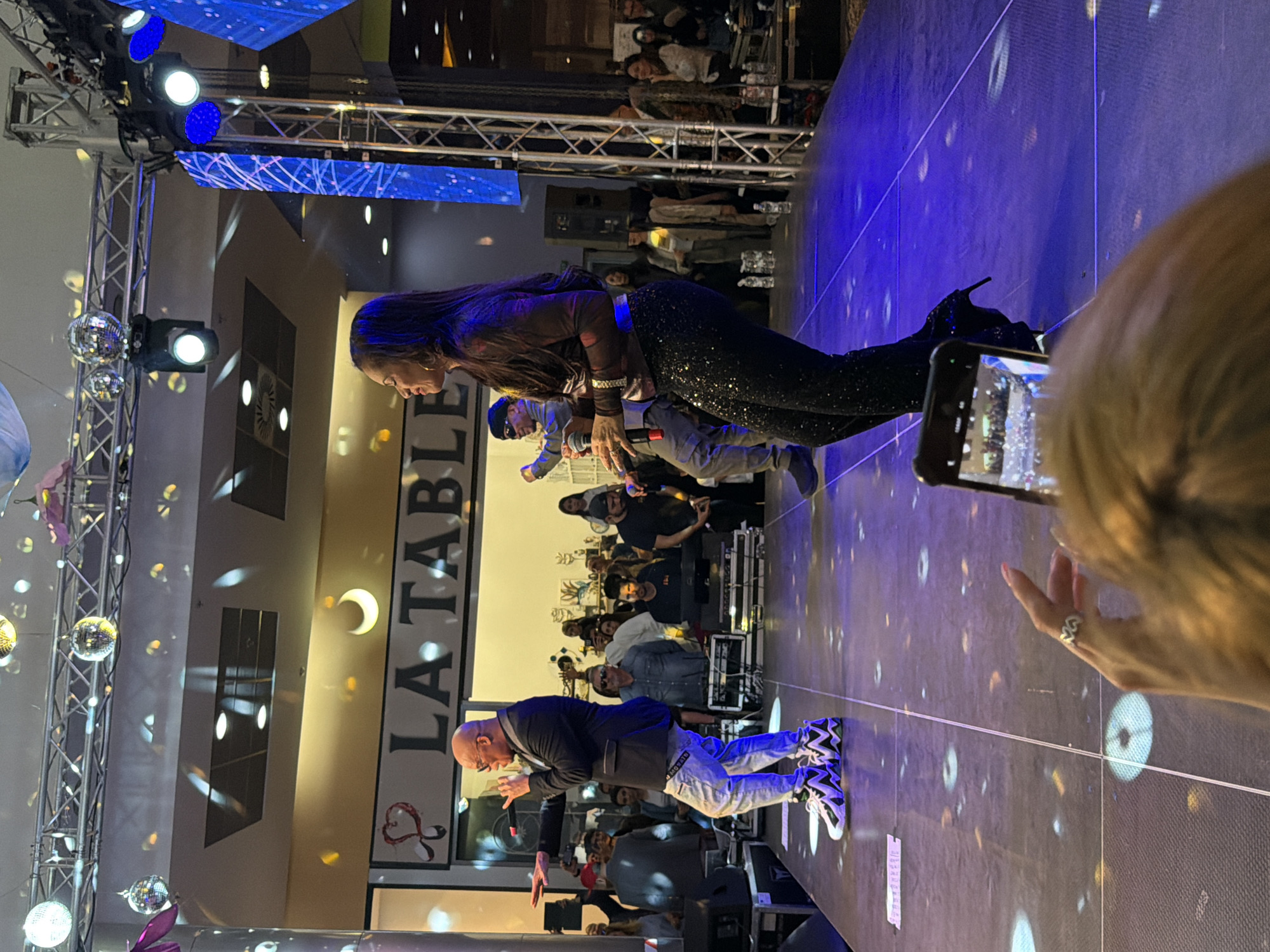 This media file captures an energetic indoor event, likely a performance or party, set at night in Pleven, Bulgaria. The image, originally rotated, shows a brightly lit stage where several individuals are performing.
In the foreground on the stage, a woman with long dark hair, wearing a dark, slightly sheer top and sparkling black pants, is visible with a microphone. To her left, a man in a dark jacket, light-colored jeans with visible branding, and distinctive black, white, and blue/purple sneakers is actively performing, also holding a microphone. Another man in a light blue shirt and cap is partially visible between them, also holding a microphone.
The stage is equipped with metal truss structures supporting spotlights and stage lights that cast blue and white illumination. Large blue screens behind the performers display abstract patterns or lights. Multiple disco balls hang from the ceiling, reflecting light and creating scattered patterns on the stage and walls.
Off-stage, a large audience is visible in the background, some standing and others seated at tables, watching the performance. In the immediate foreground, part of a person's head and hand are visible, holding a smartphone to record the event. This person has light hair and wears a ring on their finger.
A prominent vertical sign reads "LA TABLE" with a crescent moon symbol above it and what appears to be a stylized heart or intertwining symbol below. The overall atmosphere suggests a lively entertainment event.