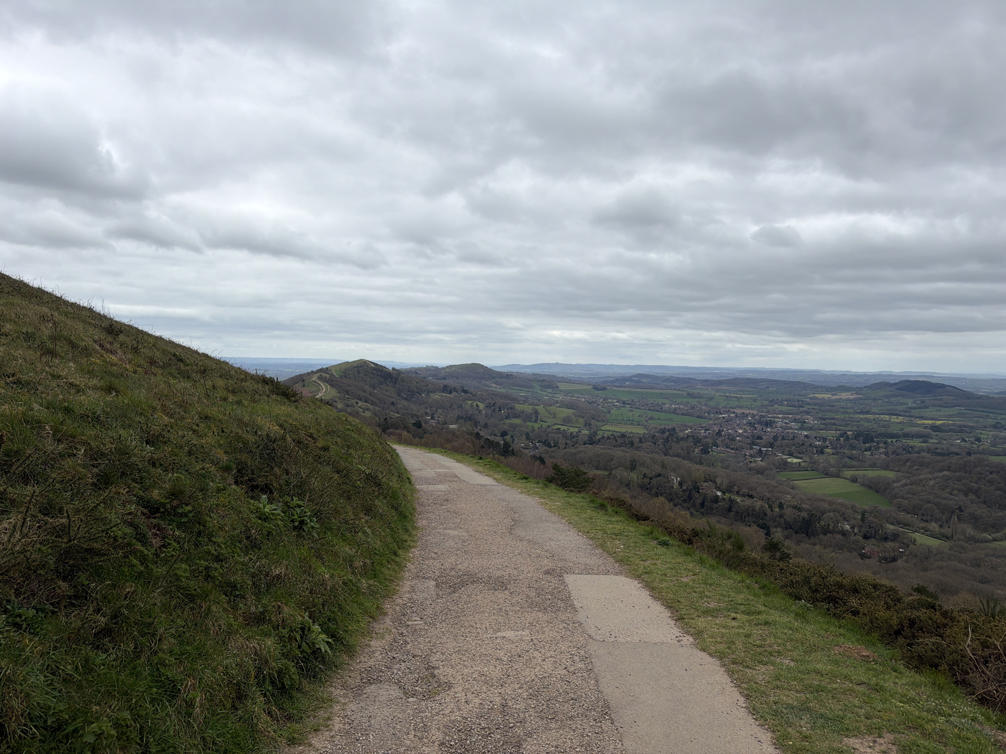 The image depicts a rural scene in the Malvern Hills, United Kingdom, under a cloudy sky. A gravel and concrete pathway stretches into the distance, winding along a hillside covered with grass and sparse vegetation on the left. The path offers a wide-angle view of a landscape comprised of rolling hills, wooded areas, and fields. A town or village is visible in the distance. No people or animals are visible in the foreground or midground.