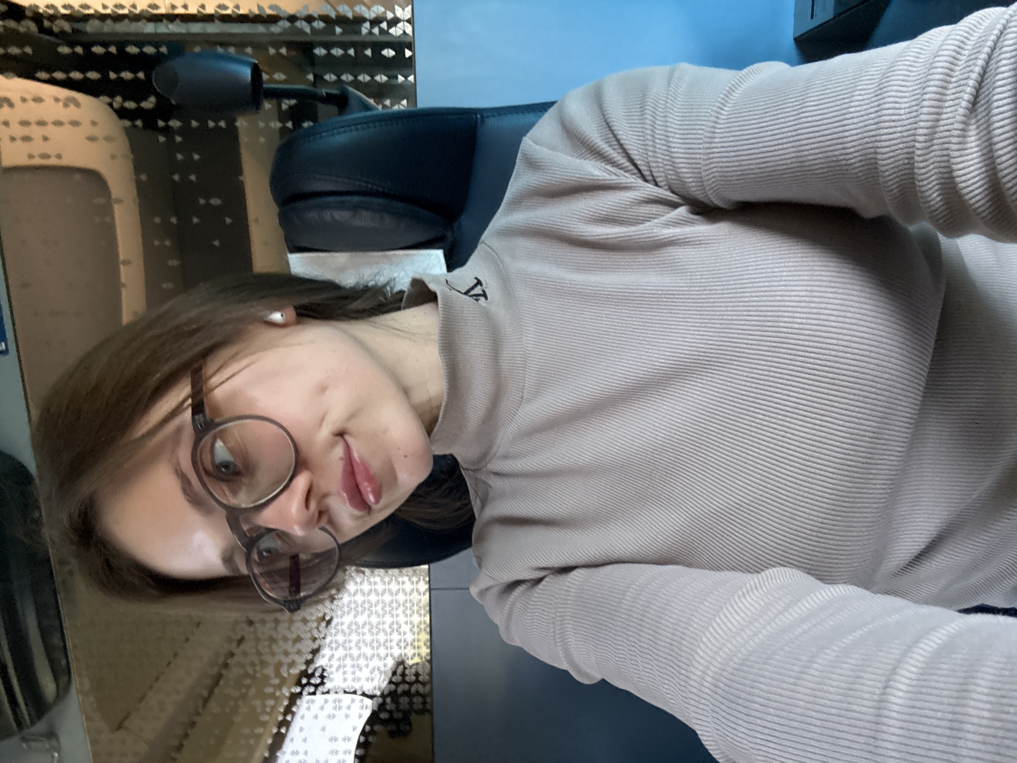 A young adult female is the central subject of the image, captured in what appears to be a selfie. The image is oriented sideways, with her head tilted to the right. She has dark brown hair, worn down, and is wearing large, round-framed glasses. A small, white stud earring is visible in her right ear. Her expression is a subtle, closed-lip smile or smirk, and she is looking directly at the camera.
She is dressed in a light beige or light brown, ribbed, long-sleeved turtleneck top. On the collar of her top, there is a small, dark embroidered logo that resembles a stylized "VT" or "LV".
Behind her, a dark-colored, possibly leather, office-style chair is visible. To the left and behind her head, a part of a pair of black headphones can be seen. The background features a wall or partition with intricate, repeating geometric patterns (small squares or diamonds) in light and dark tones. Above this patterned section, a solid blue area is visible, possibly another wall or a screen. The lighting is soft, suggesting an indoor setting.
The scene takes place indoors, likely in a personal or workspace environment, indicated by the presence of an office chair and headphones. No specific time of day is explicitly indicated, but the lighting suggests daytime.