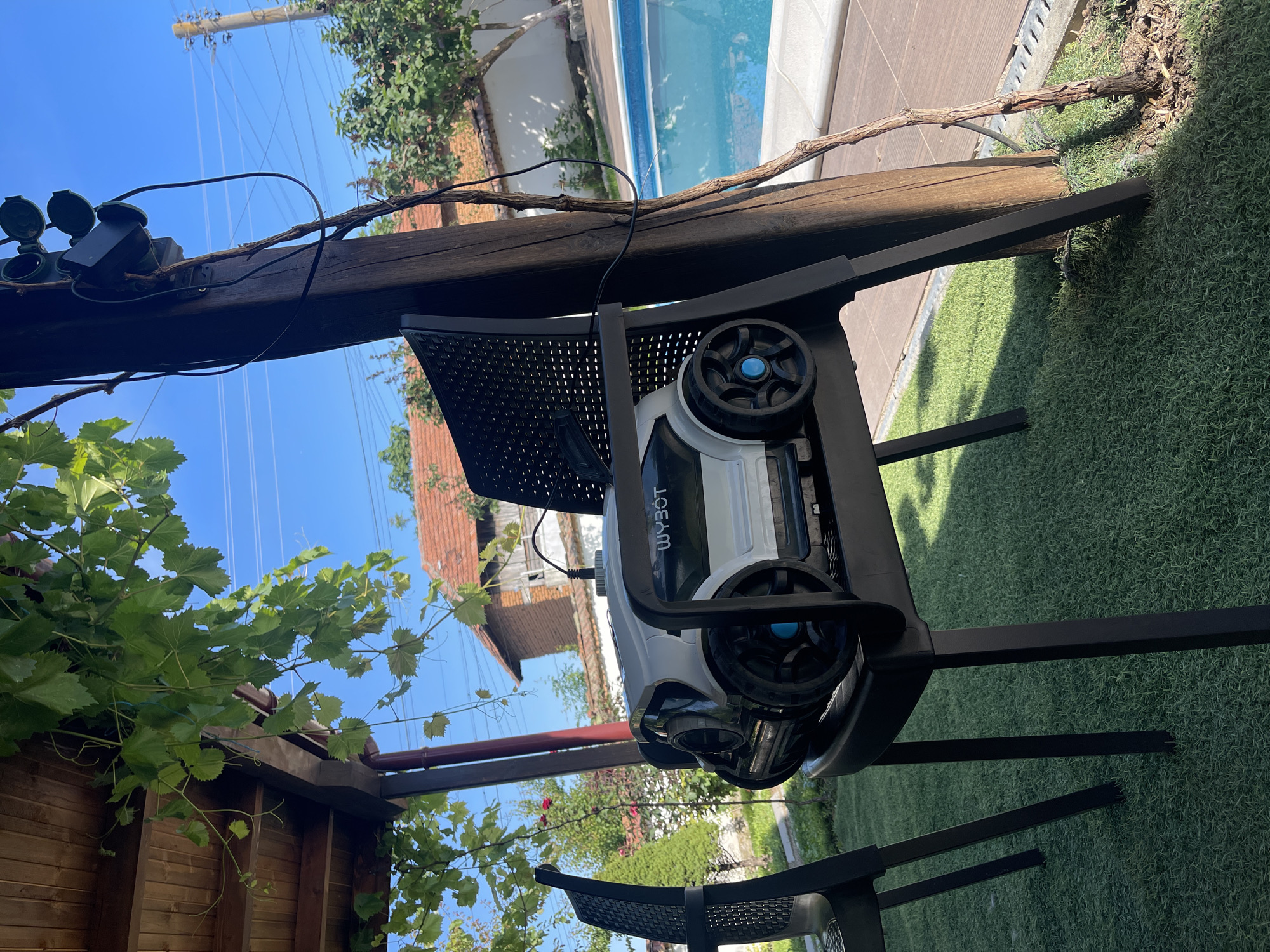 A white and black robotic lawnmower, identified by the "WYBOT" logo, sits on a black plastic chair under a wooden pergola. The chair and lawnmower are positioned in a yard with artificial grass, adjacent to a swimming pool. In the background, a building with a red tile roof and trees are visible. The scene appears to be outdoors in daylight, with a clear blue sky. This location is in Totleben, Bulgaria.