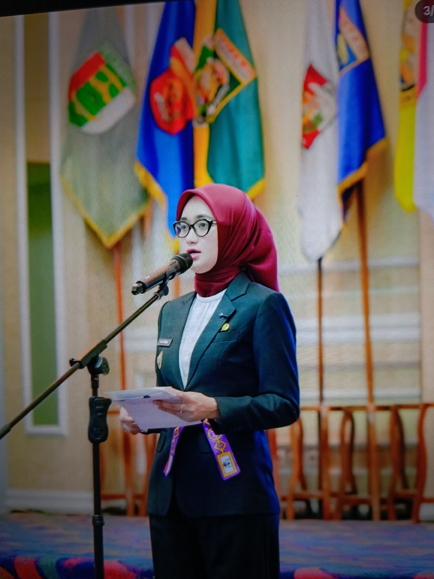 A woman wearing a red hijab and glasses is speaking at a podium in front of several flags. She is wearing a dark suit jacket over a white shirt and has a lanyard with a purple and gold badge around her neck. The flags are colorful and appear to be official or ceremonial. The room has wooden chairs in the background and a patterned carpet on the floor. The lighting suggests an indoor event. The image is a photograph taken from a slightly low angle.
