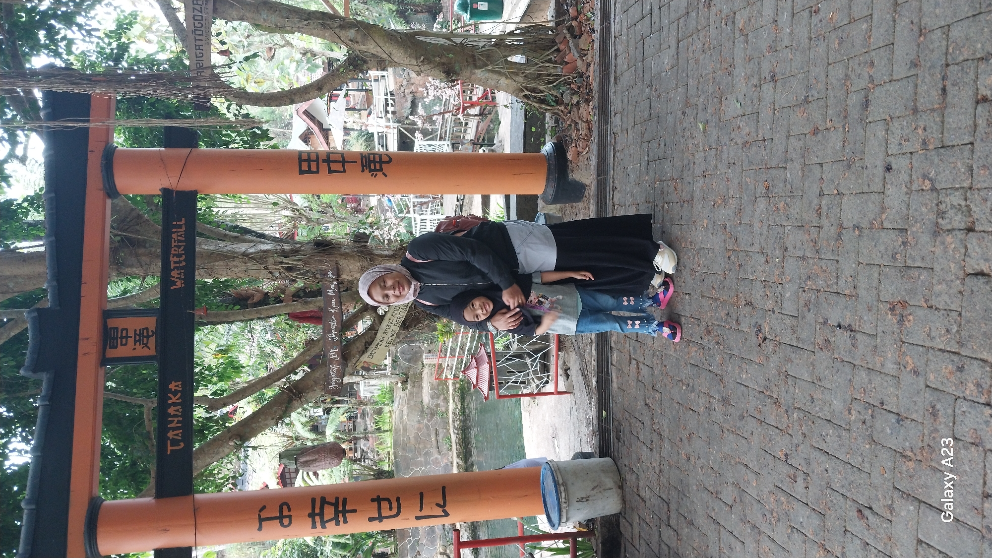 The image shows a woman and a young girl standing in front of an orange torii gate with Japanese inscriptions. The gate is located outdoors, with trees and greenery in the background, suggesting a natural setting. The ground is paved with brick. The woman is wearing a black jacket, a light-colored skirt, and a pink hijab. The girl is wearing a grey t-shirt and blue jeans. They are both smiling. The torii gate has Japanese characters painted on it, one column on the left and one on the right. Above the arch, there is a horizontal sign with the text "TANAKA WATERFALL". The overall scene appears to be a tourist attraction. It is identified as being in Bangelan, Indonesia.