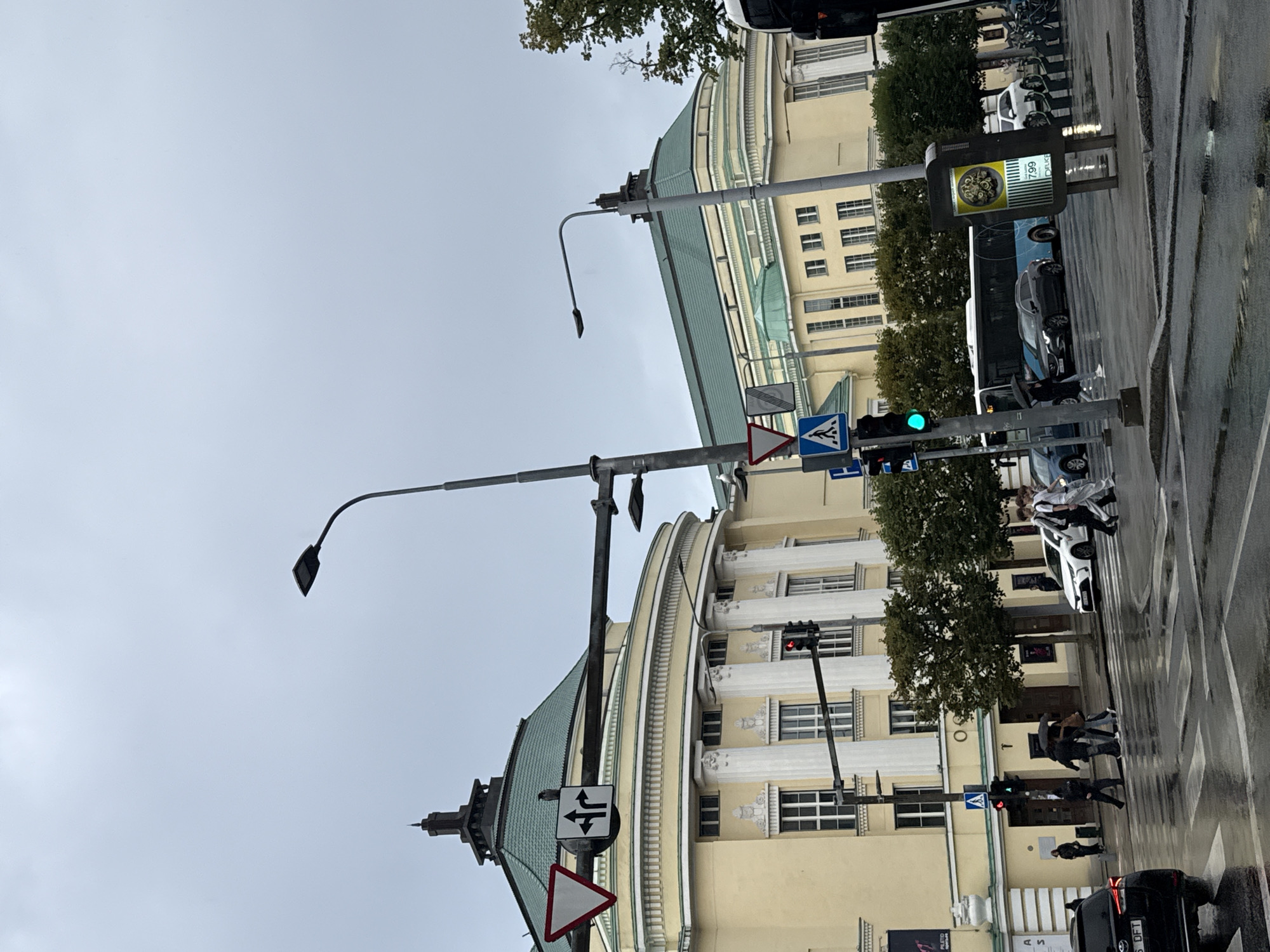The image shows a street scene in Tallinn, Estonia, likely in front of the Estonian National Opera building. The weather is overcast, and the street is wet, indicating recent rain.
In the foreground, a black SUV is partially visible on the left. To the right, pedestrians are crossing a wet street at a crosswalk. Some are carrying umbrellas. Traffic lights are visible, with one showing a green light.
The background features a light yellow building with white columns and a distinctive green roof, consistent with the architecture of the Estonian National Opera. A blue bus is also visible in the street. Traffic signs indicating road directions and pedestrian crossings are present. The overall scene depicts a typical urban environment on a damp day.