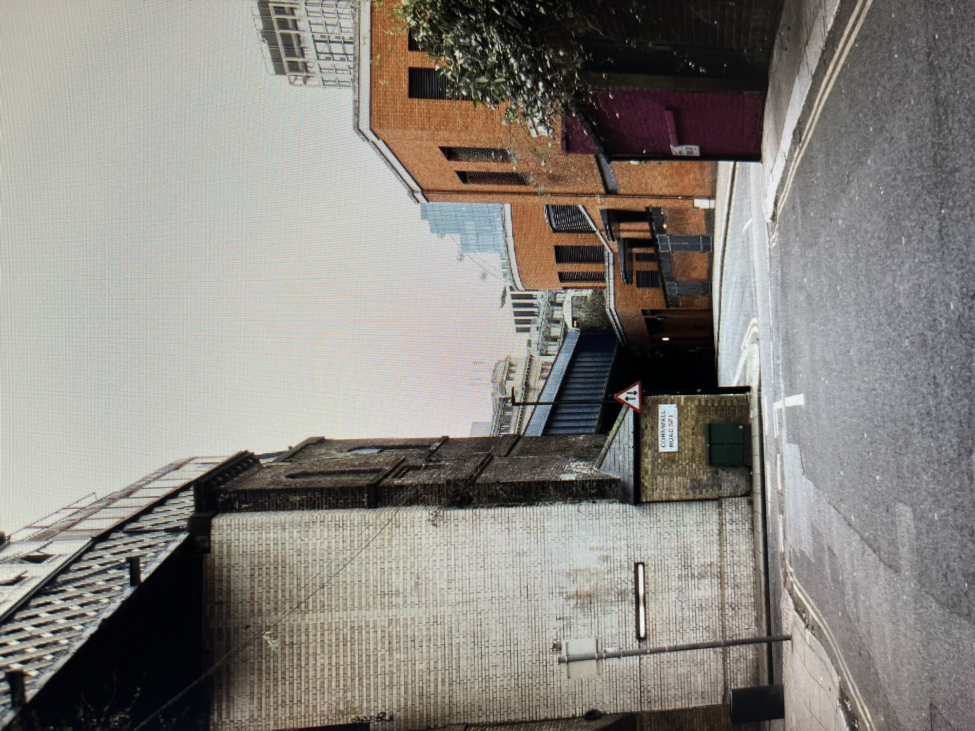 This is an eye-level, medium shot of a narrow street with an overhead railway bridge. The street is paved with asphalt and has white road markings. On the left, a large, weathered brick building with a white-washed lower section dominates the foreground. A street lamp is attached to the wall. Next to this building is a smaller, brick structure with a green door and a sign that reads "CORNWALL ROAD SE1". A red traffic sign with a white border and a white symbol indicating two-way traffic is affixed to the wall of this smaller structure.
The street curves gently to the right. On the right side of the street, a modern orange brick building with dark-shuttered windows is visible. Adjacent to it is a dark purple door. Further in the background, a glimpse of taller buildings, possibly skyscrapers, can be seen. The sky is overcast. The scene is devoid of people.