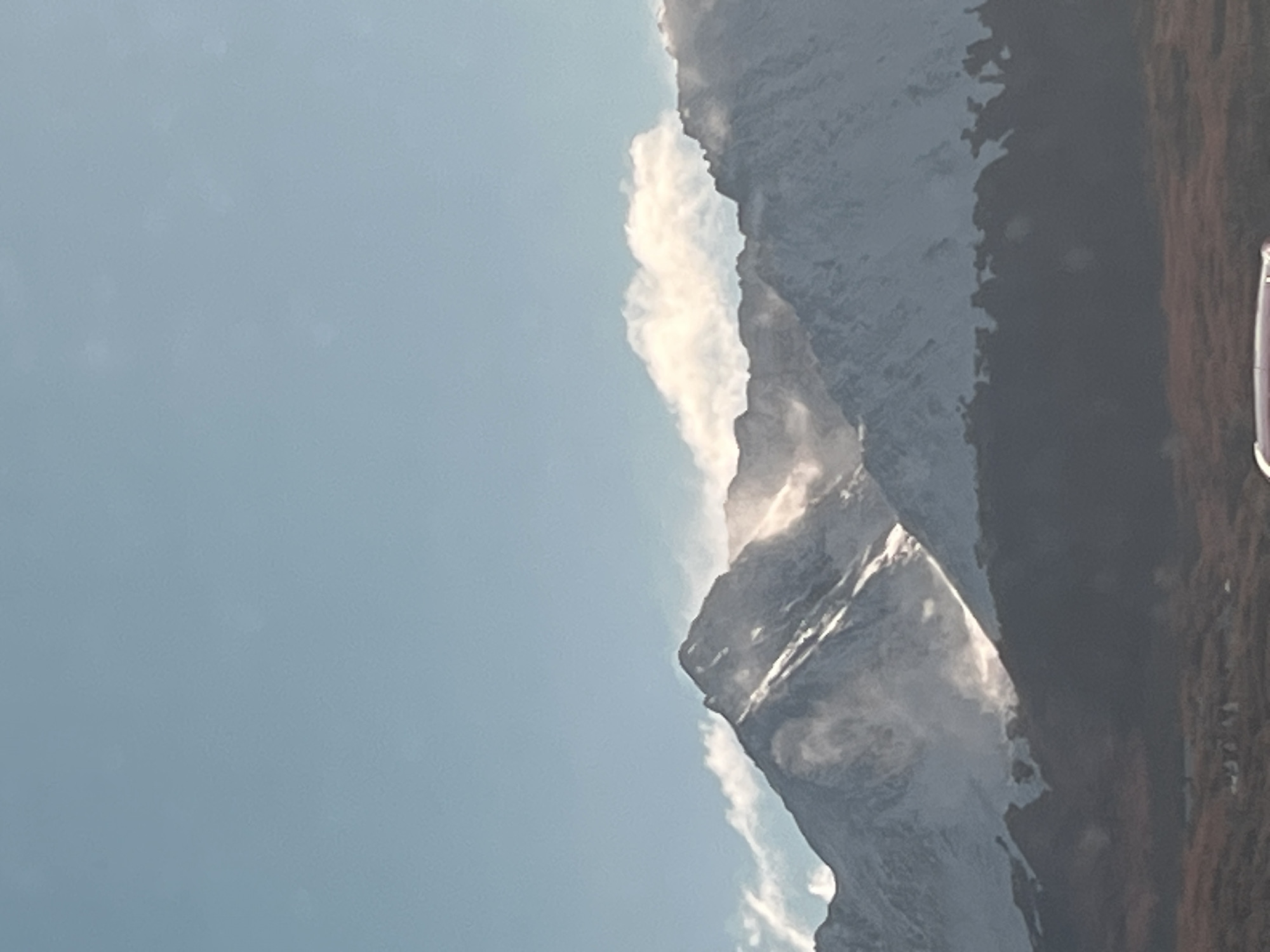 The image depicts a mountainside partially covered with snow, possibly located in the vicinity of Ushuaia, Argentina. The mountain is rugged, with sharp peaks and ridges, and a steep slope. Some clouds are visible near the top of the mountain. The sky is light blue. The edge of a window is visible at the right of the picture.