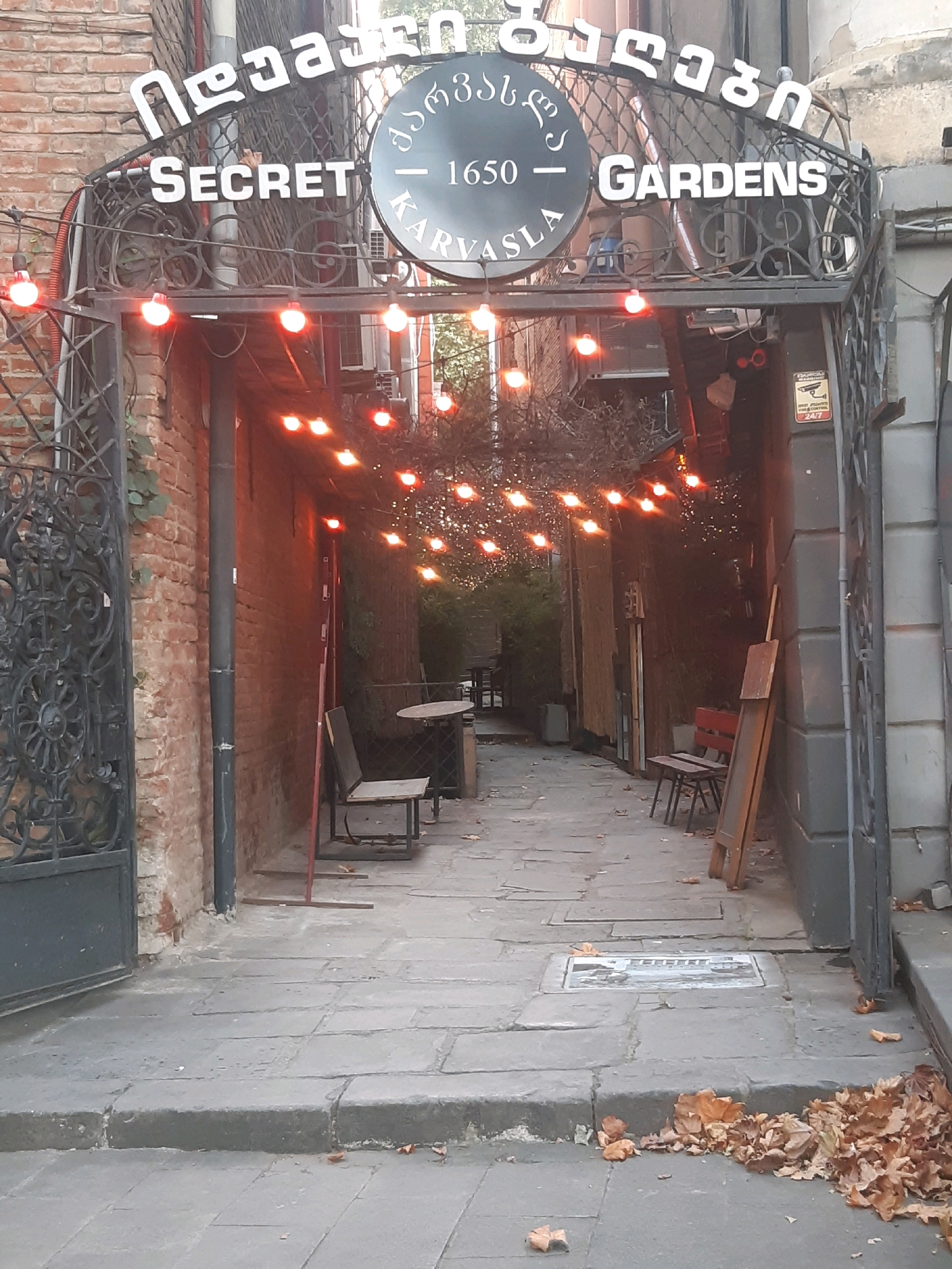 The image shows the entrance to "Secret Gardens Karvasla" in Tbilisi, Georgia. The entrance is an archway decorated with lights and a sign with the establishment's name in English and Georgian script. The archway leads to a narrow pathway with brick walls on either side, and outdoor seating areas. The pathway is paved with stone slabs, and fallen leaves are scattered on the ground, indicating it is autumn. The scene is a view looking into the establishment from the street.