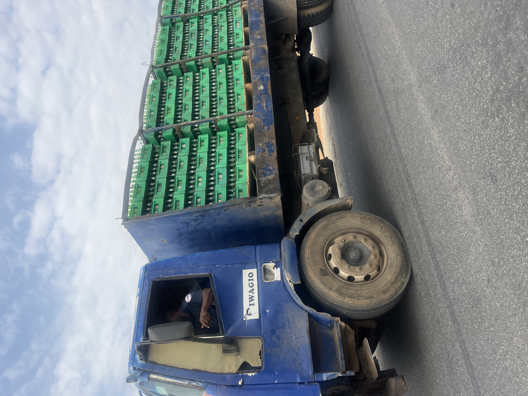 A blue, well-used flatbed truck is captured on a paved asphalt road in Karum, Nigeria, under a bright, cloudy daytime sky. The truck's flatbed is heavily loaded with numerous green plastic crates, stacked several layers high and supported by metal frameworks. Each crate appears to contain multiple glass bottles, likely for beverages.
A male driver is visible in the truck's cab, leaning out of the open window with his right arm resting on the window frame. He is wearing a dark shirt. The truck's cab shows signs of wear and tear, including visible dirt and some damage or missing panels on the passenger side. The tires appear robust, suited for heavy transport.
The setting is an outdoor, clear day, with scattered white clouds in the sky. The road is a dark gray asphalt surface. On the side of the truck's cab, a sticker or painted text clearly displays the word "IWAGIO". The overall scene depicts a commercial vehicle engaged in the transport of goods.