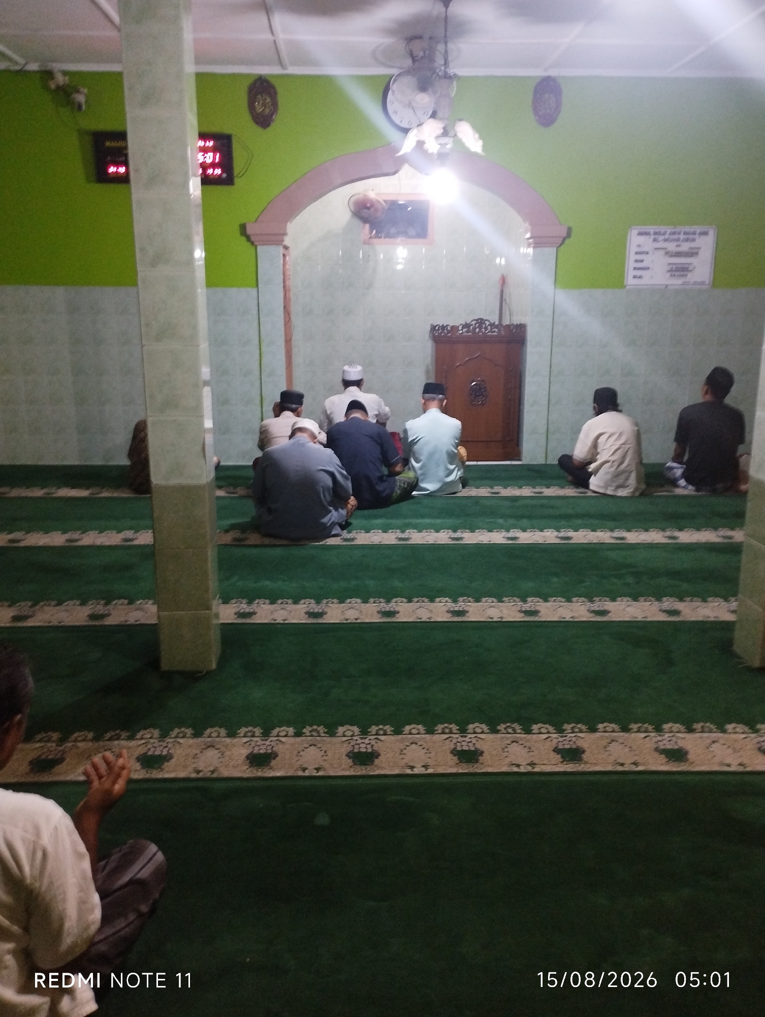 The image depicts the interior of a mosque in Karawang, Indonesia. A group of men are seated on a green carpet, facing away from the camera, in prayer. Some men wear white skullcaps, while others wear traditional Islamic attire. A wooden pulpit is visible in the background. The walls are painted green and decorated with Islamic calligraphy. A digital clock displaying the time "05:01" and the date "15/08/2026" is present on the wall. The lighting appears to be artificial, casting a warm glow on the scene. The overall atmosphere is one of solemnity and devotion.