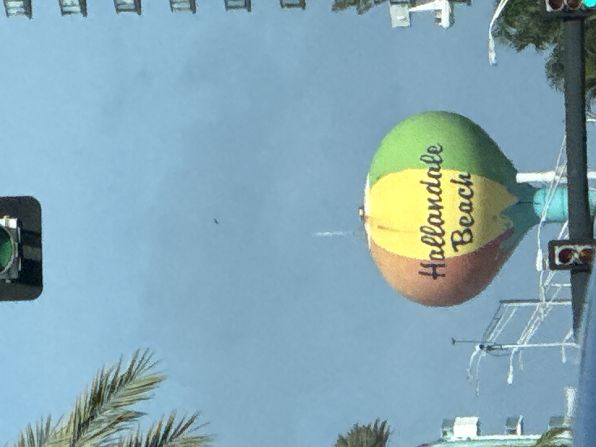 The image shows a water tower painted to resemble a hot air balloon, with the words "Hallandale Beach" written on it. The water tower is located in Hallandale Beach, Florida, United States of America. The sky is blue, and there are some palm trees and a traffic light in the foreground. A tall building is visible in the upper left corner of the image.