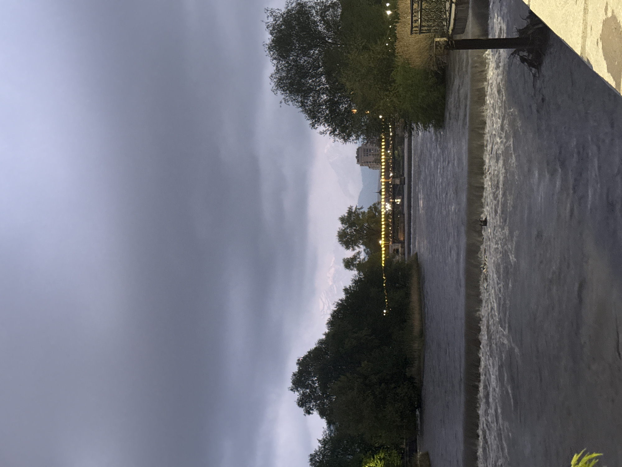 The image shows a river flowing in the city of Komgaron, Russia. The view includes a cityscape faintly visible in the distance, framed by dark clouds. There are lights along the riverbank. Vegetation lines both sides of the river. The water flows from a higher level to a lower level. There are no people visible in the image.