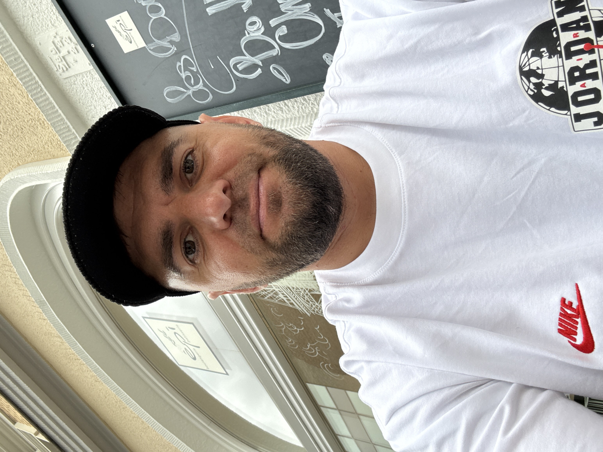 The image is a selfie taken by a man with a beard and a black baseball cap, wearing a white t-shirt with red Nike and Jordan logos. He is positioned in front of a building with an arched window and a chalkboard sign. The sign, written in white script, appears to be a menu or advertisement. Behind the man, a building facade with beige textured walls and white architectural details is visible. To the left of the man, a sign with "épi" and "boulangerie patisserie" is displayed. The man is looking directly at the camera, and his expression is neutral. The overall setting appears to be an outdoor cafe or shopfront in Bonn, Germany.
