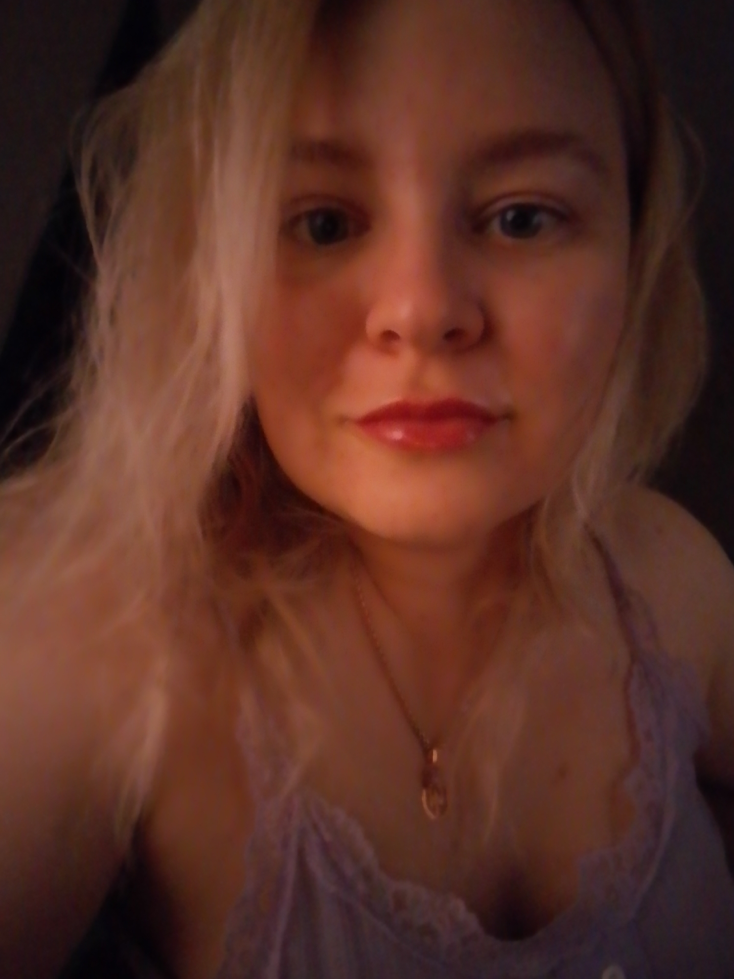The image is a selfie of a fair-skinned woman with blonde, wavy hair. She has blue eyes and is wearing red lip gloss. She is wearing a light purple camisole with lace trim and a gold necklace with a pendant. The background is dark and out of focus. The image provides no information about the city of Izhevsk, Russia.