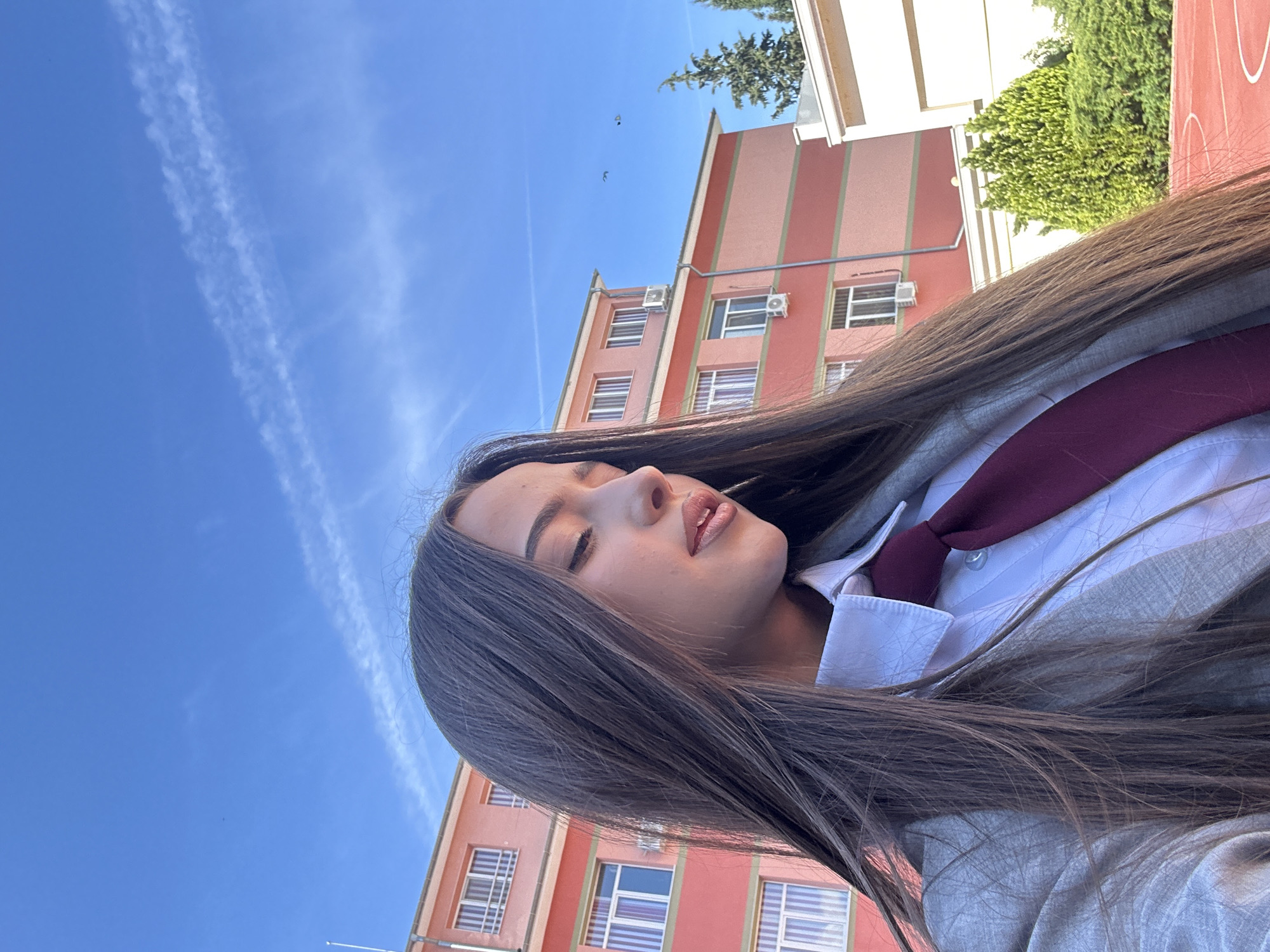 A young woman with long, dark hair is seen from a low-angle perspective, posing outdoors with her eyes partially closed. She is wearing a white shirt, a maroon tie, and a grey blazer. The background features a light blue sky with contrails and a peach-colored building with rows of windows and what appear to be air conditioning units. Green foliage can be seen near the building. The photograph appears to have been taken in an urban environment, possibly a school or campus setting.