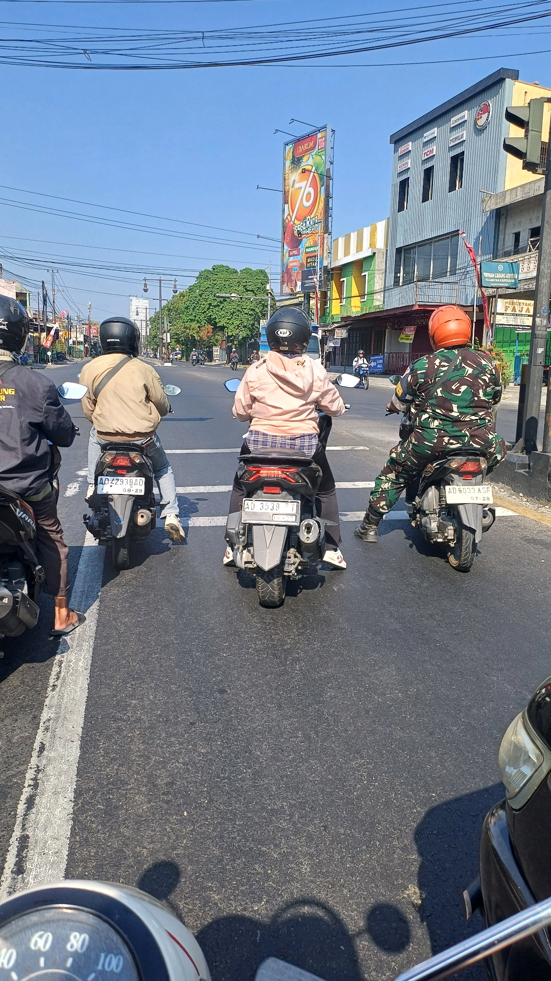 In Pucangan, Indonesia, on a sunny day, four motorcyclists are stopped at a traffic intersection. The motorcyclists are all wearing helmets, and their backs are to the camera. The first motorcyclist on the left is wearing a black jacket and brown pants. The second motorcyclist is wearing a tan jacket and blue jeans. The third motorcyclist is wearing a pink hooded jacket and plaid shorts. The fourth motorcyclist on the right is wearing a camouflage military uniform. All four motorcycles are parked in a single file line, facing forward. The road is paved and has white lane markings. In the background, there are buildings, trees, and a large billboard. The billboard has a picture of a lion and the words "DIARUM" and "76." The traffic light is red.
