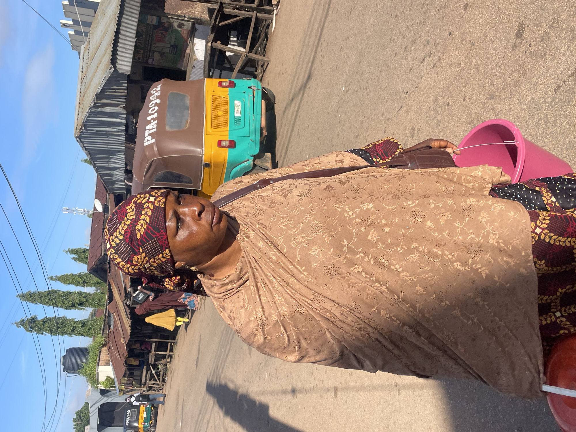 The image depicts a street scene in Jos, Nigeria, likely near a market. A woman is in the foreground, walking towards the viewer. She is wearing a brown headscarf with a red and gold pattern, and a flowing brown garment with a subtle floral print. Her lower body is adorned with a colorful patterned fabric. She carries a brown bag over her shoulder and a pink bucket in her right hand.
In the background, a yellow and green auto-rickshaw, possibly a commercial vehicle, is parked on the right side of the road. The rickshaw has the license plate "PTA-10942" visible. To the left of the rickshaw, several market stalls with corrugated iron roofs are set up along the street. People can be seen moving around in front of these stalls. A tall black water tank is visible on a building behind the stalls, and several tall, slender trees line the edge of the road. Power lines crisscross the sky. The road surface appears to be paved with asphalt, and the scene is illuminated by bright sunlight, casting shadows on the ground. The overall impression is one of a bustling urban environment in Nigeria.
