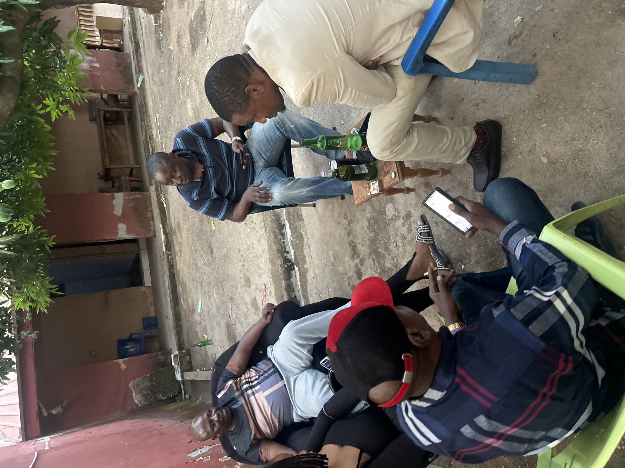 In an outdoor setting in Jos, Nigeria, several individuals are gathered. At least six people are visible, mostly male. One man, wearing a red baseball cap, is seated, while another individual is using a mobile phone. Several empty green beer bottles are on a small table. The environment appears to be a concrete courtyard. A red wall is visible in the background.