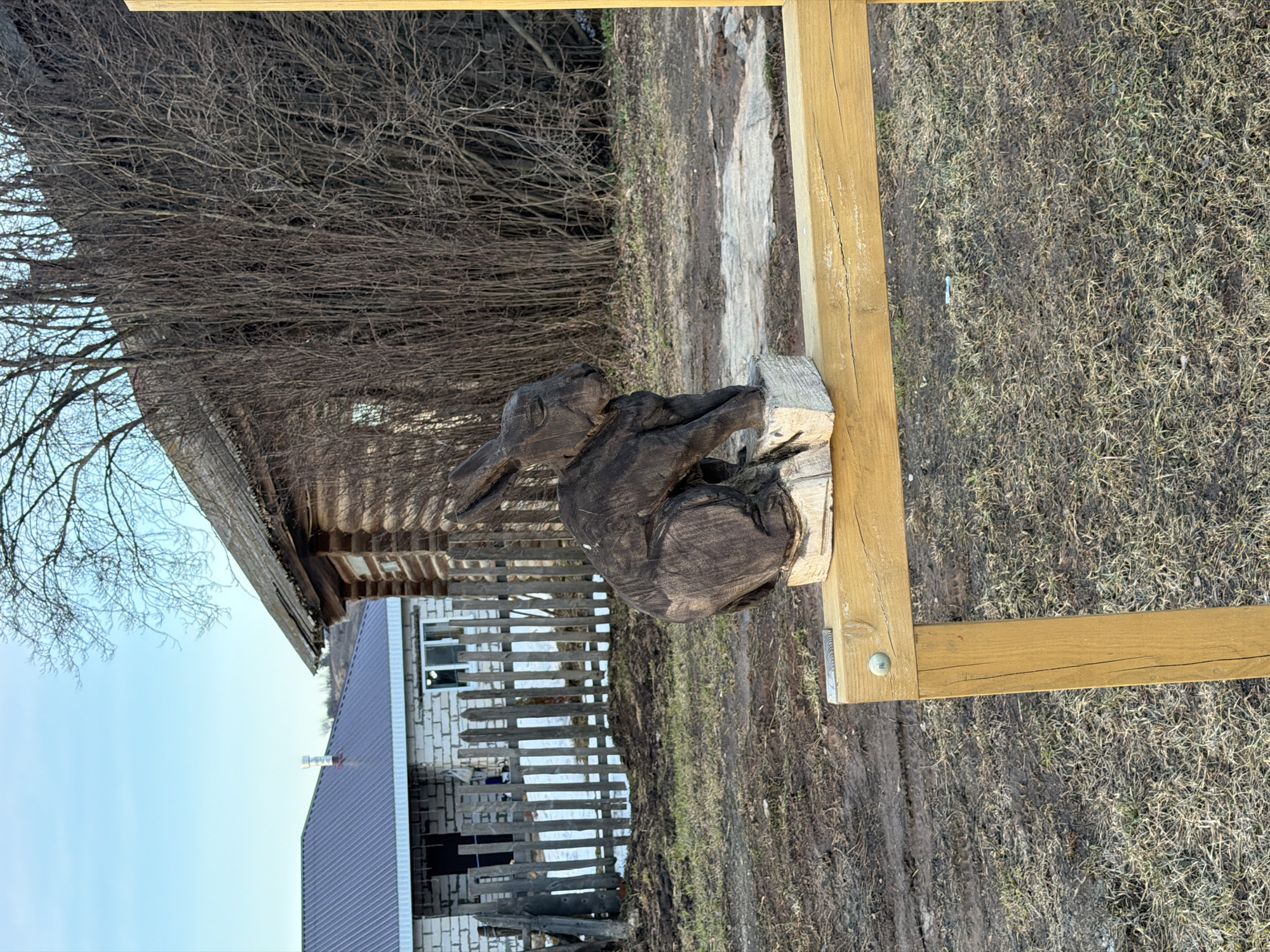 The image depicts a rural scene in Дулесово, Russia. A carved wooden rabbit sculpture rests on a wooden block, which is positioned on a wooden frame. In the background, there's a rustic wooden building with bare branches obscuring parts of it. Adjacent to the wooden building is a brick structure with a grey roof and a wooden fence in front. The ground is covered in dry grass and dirt.