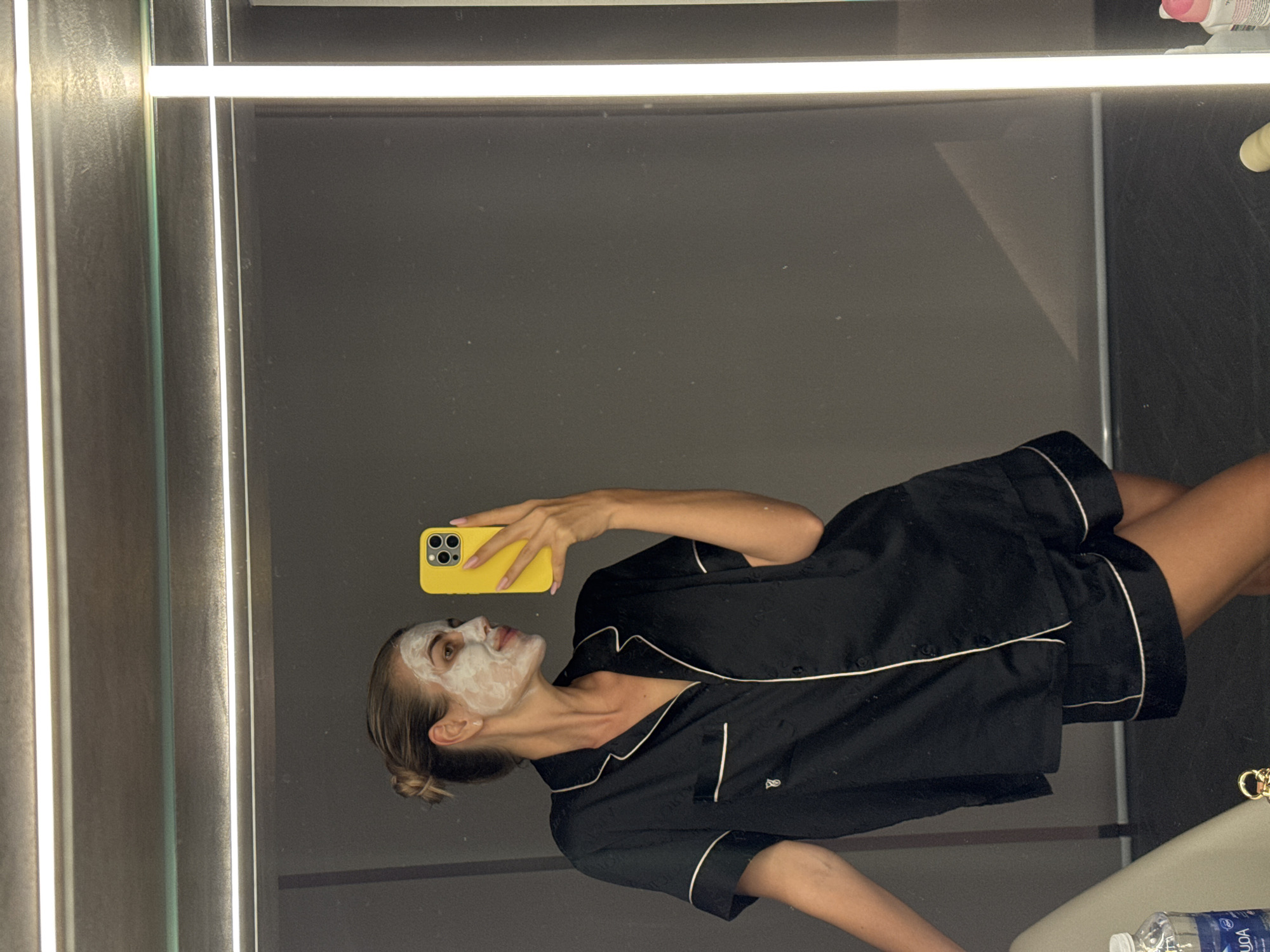 A woman in a black pajama set with white piping is taking a selfie in front of a mirror. She has a white face mask on and is holding a yellow phone. A water bottle and various toiletries are visible on a sink to the right. The mirror has a light at the top. The location is an unknown bathroom in an unknown city.
