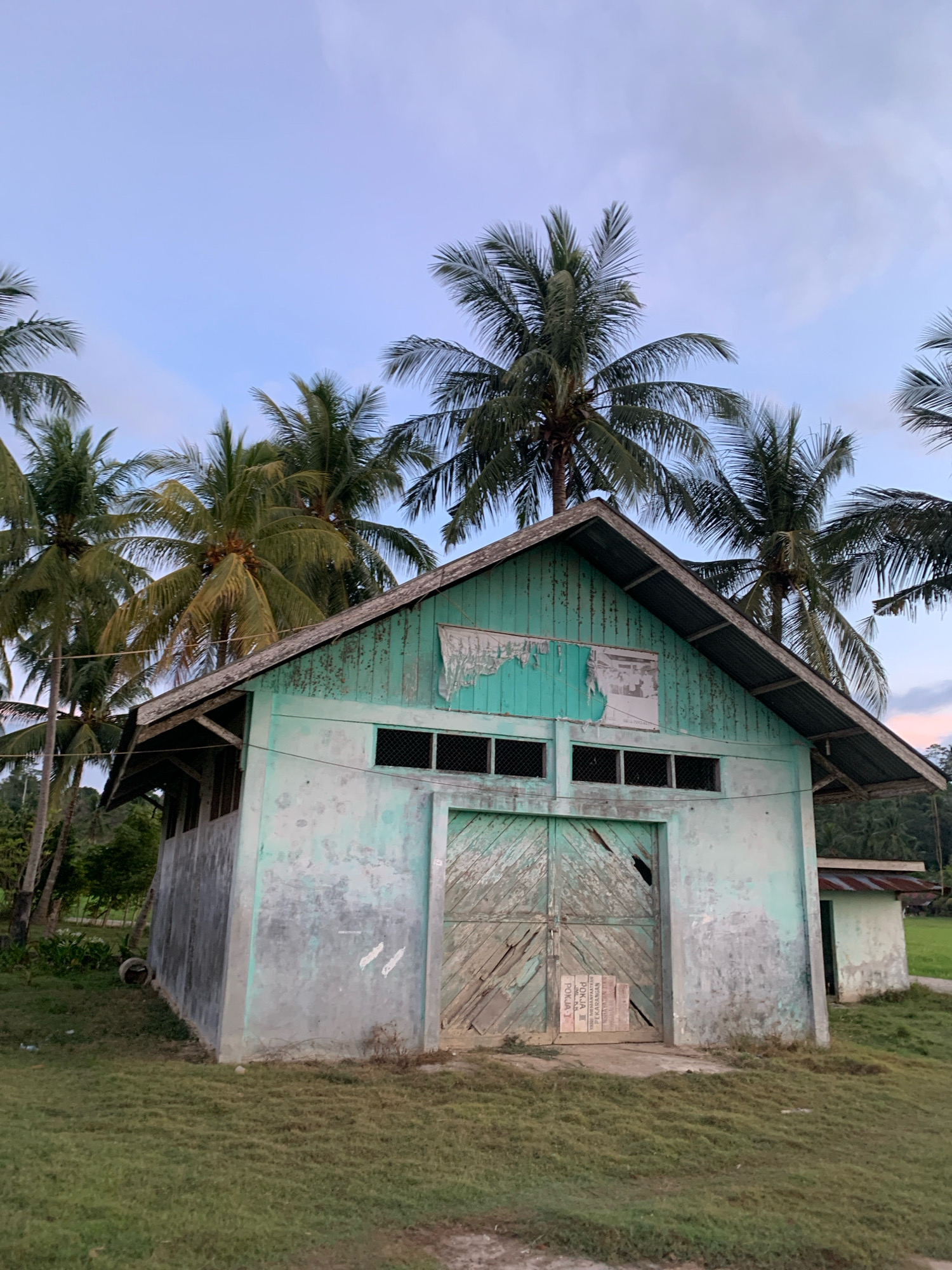 The image depicts an abandoned building with peeling green paint, located in the city of Palohawe, Indonesia. The structure features a large, weathered wooden door and several windows with wire mesh. Palm trees surround the building, and the sky is a pale blue with wispy clouds. The surrounding area is covered in overgrown grass. A smaller, similarly dilapidated structure is visible to the right of the main building. The scene suggests a rural or neglected environment.