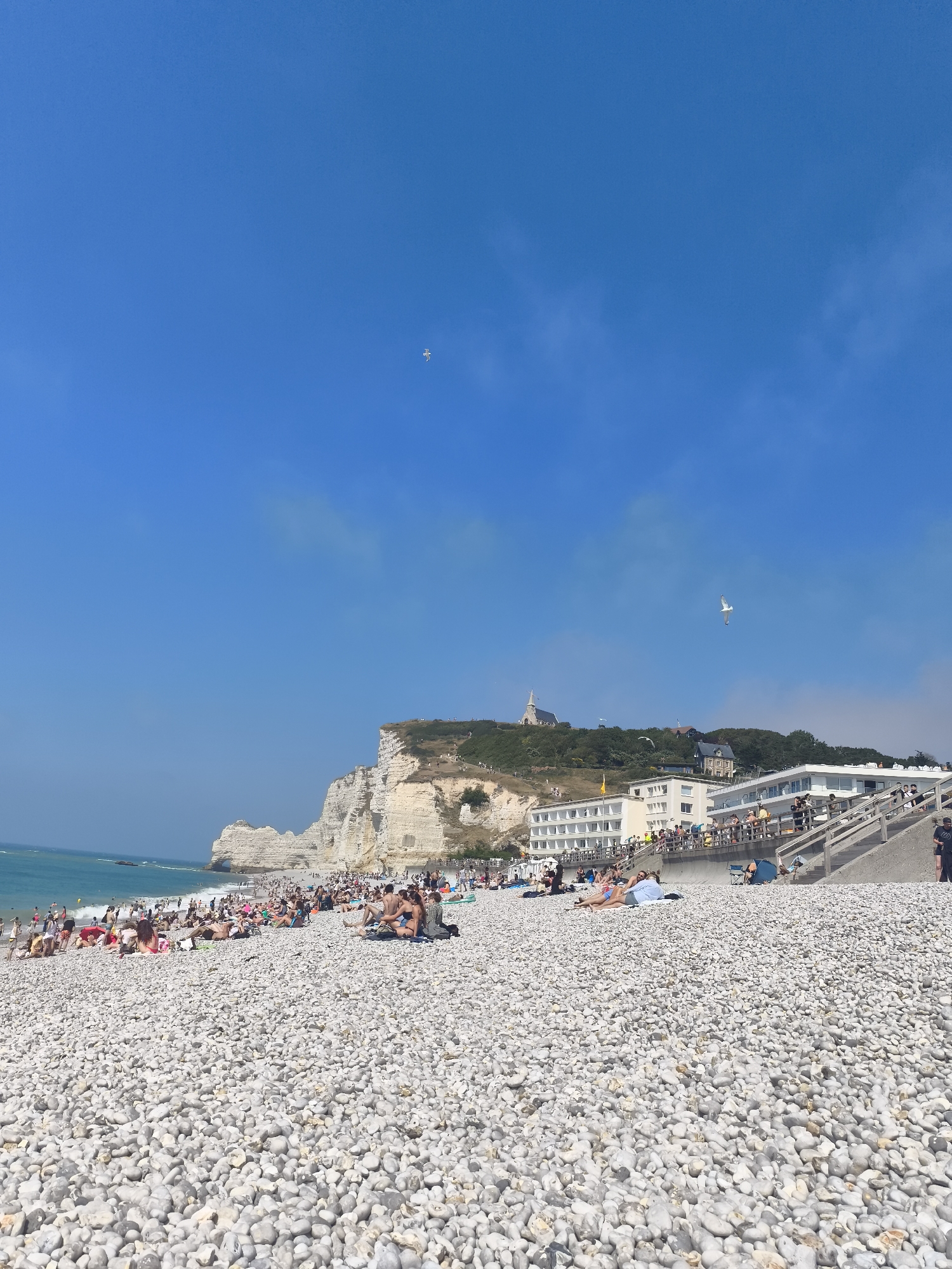 The image depicts a crowded pebble beach in Étretat, France, under a clear blue sky with scattered clouds. In the foreground, white and grey pebbles fill the majority of the frame. Numerous people are visible on the beach, some sunbathing on towels and blankets, others sitting or walking. The middle ground shows a white building with multiple floors and a balcony structure, teeming with people. Beyond the building, lush green hills rise, topped with trees and a prominent church steeple. To the left, the iconic white cliffs of Étretat, featuring a natural archway, rise majestically from the turquoise ocean. The water is calm and stretches to the horizon. Two birds are visible in the sky, one higher up and slightly to the left, and another, a seagull, is in flight closer to the buildings. The scene conveys a lively summer atmosphere on the French coast.