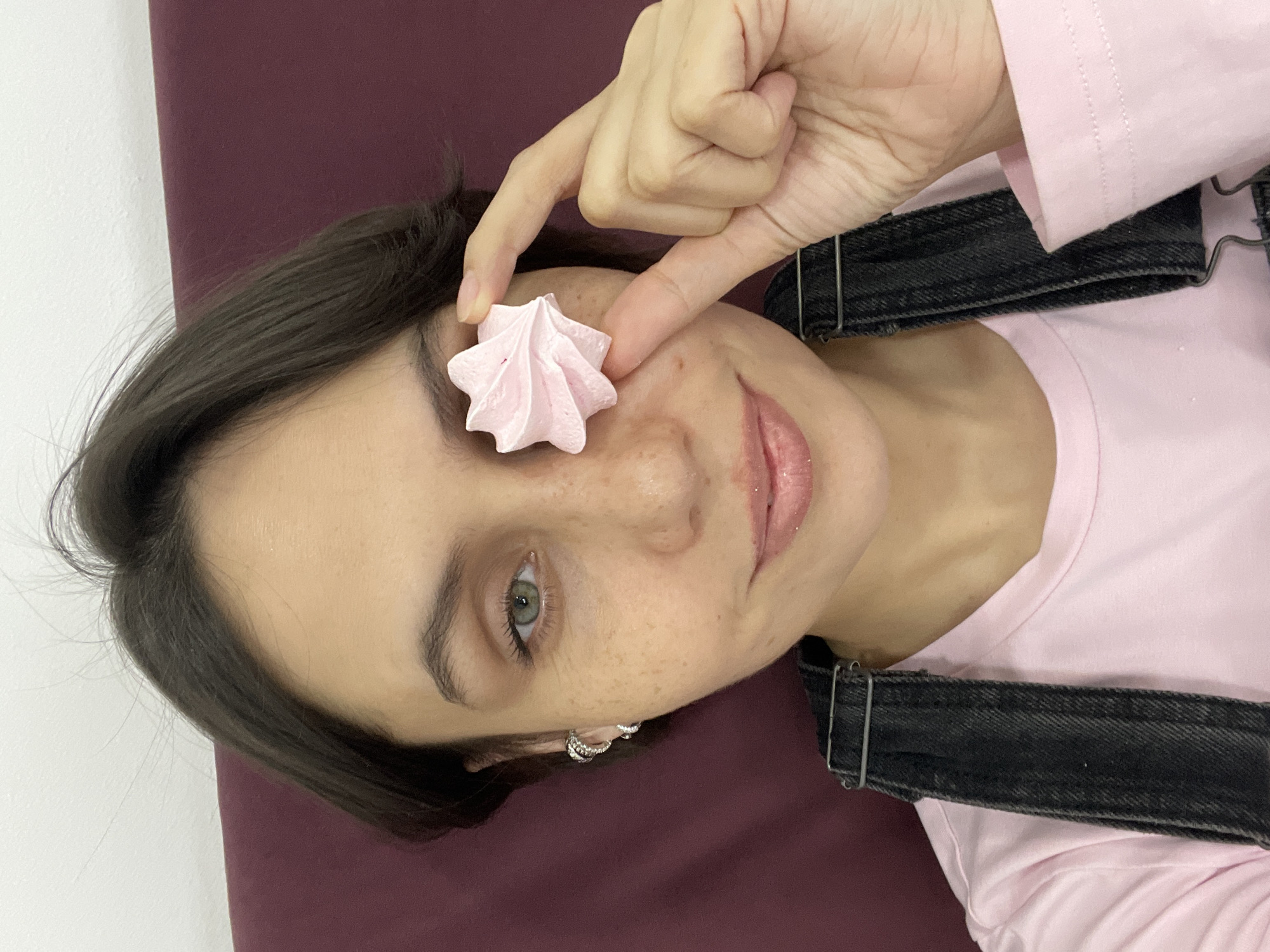 A young woman with dark brown hair, fair skin, and light greenish-grey eyes is featured in a close-up, diagonal selfie taken indoors in Izhevsk, Russia. She has freckles across her nose and cheeks and wears a light pink lip gloss. She gazes directly at the camera with a subtle, gentle smile.
In a playful pose, she holds a light pink, star-shaped meringue cookie between the thumb and forefinger of her right hand, placing it directly over her left eye. She is dressed in a light pink long-sleeved shirt layered under dark denim overalls, and a small silver-toned hoop earring is visible in her left ear. The background is simple, consisting of a plain white wall on the left and a solid dark purple or maroon surface behind her head and body. The lighting indicates an indoor setting. No text is visible in the image.