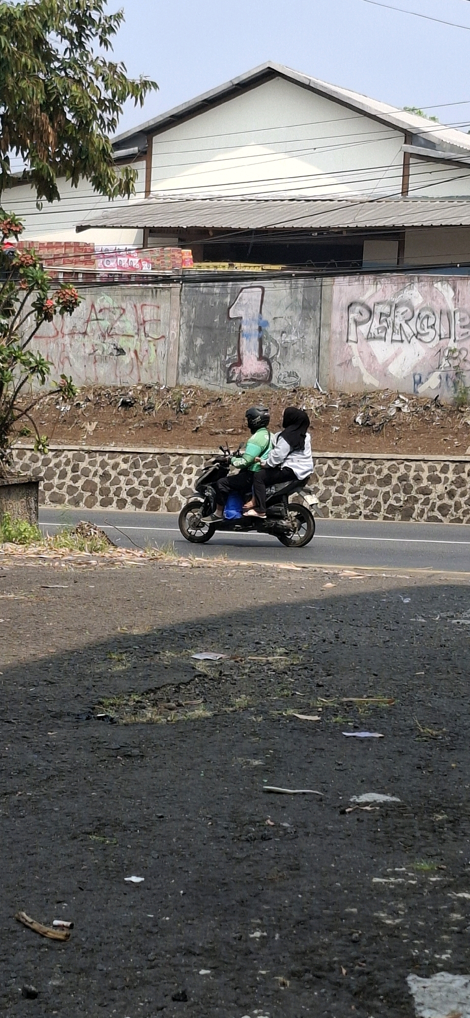 In Cilaku, Indonesia, a man and a woman are riding a motorcycle on a paved road. The man is driving, wearing a green shirt and a black helmet, while the woman is a passenger, wearing a white long-sleeved shirt and a black hijab. They are positioned side-by-side on the motorcycle. The background features a stone retaining wall and a concrete wall with graffiti, including a large red number "1" and the word "PERCIBE". Above the wall, a building with a white facade and a metal roof is visible, along with power lines and some foliage. The foreground shows a dark, rough asphalt surface with scattered debris. The scene appears to be outdoors during the daytime.