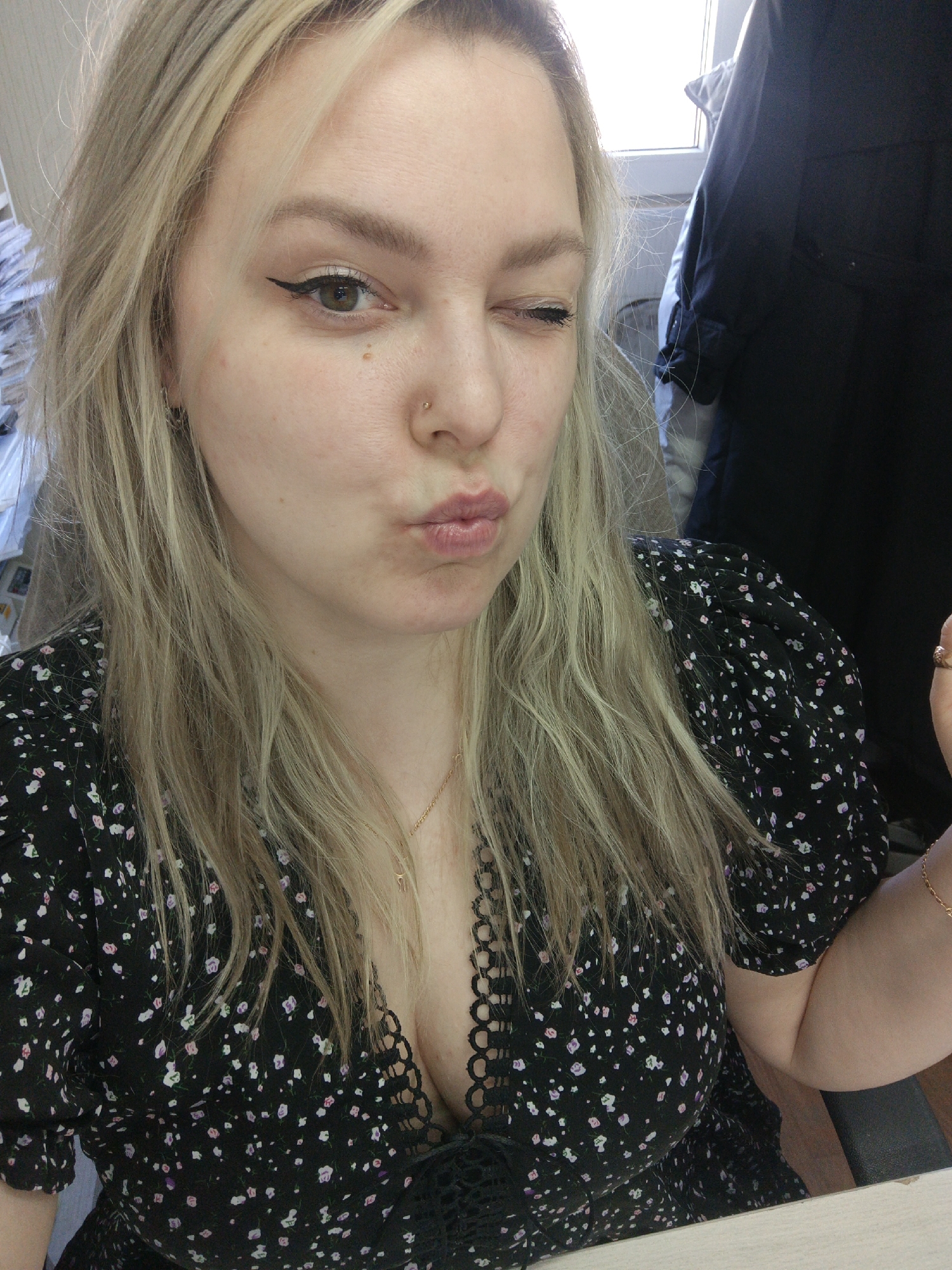 A blonde-haired woman with a nose piercing is winking at the camera. She is wearing a black dress with a floral pattern and a gold necklace. She is making a kissing face. The location is Noviy, Russia. In the background, there is a window, some papers, and a black garment hanging. She is sitting at a desk.