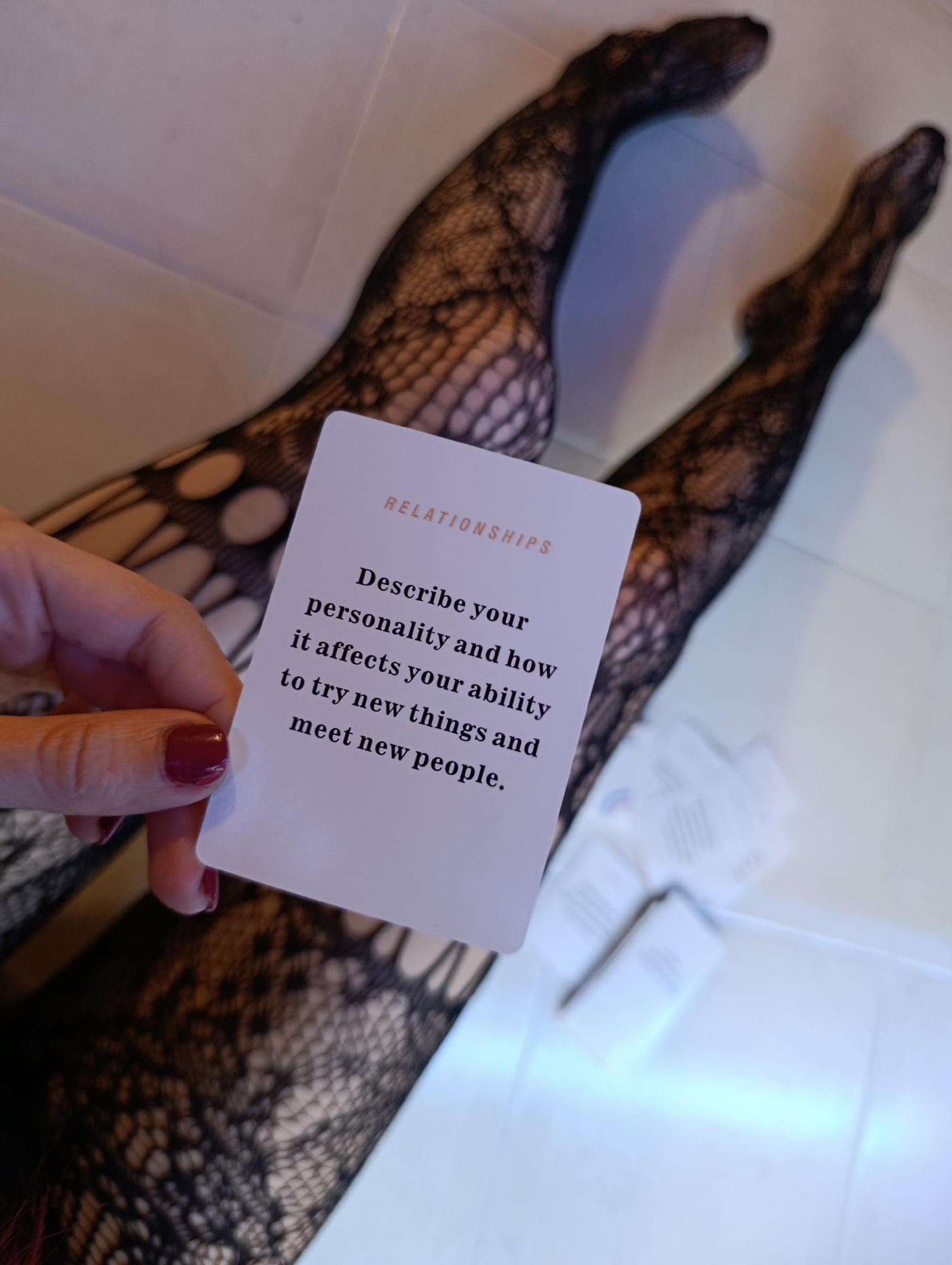 A person wearing patterned black tights lies on a white surface, holding a white card with the word "RELATIONSHIPS" in orange, followed by the prompt "Describe your personality and how it affects your ability to try new things and meet new people." A hand with red nail polish holds the card. The location appears to be indoors. The image does not provide any information to indicate the city of Svilengrad, Bulgaria.