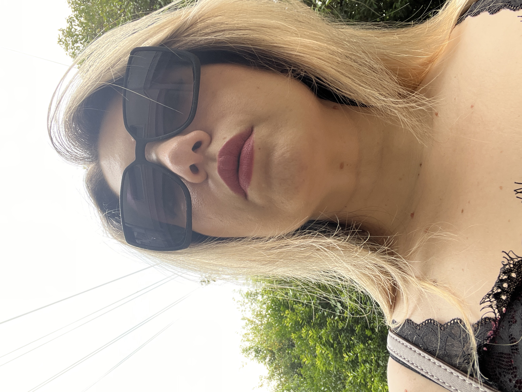 The image shows a low-angle, close-up shot of a woman wearing large, dark sunglasses and dark red lipstick. Her blonde hair is partially visible around her face, and she is wearing a black lace top. The background consists of lush green foliage and a bright sky with what appear to be overhead wires. The overall impression is that of a candid, outdoor photograph taken on a sunny day. There is no indication of the location being Sochi, Russia.