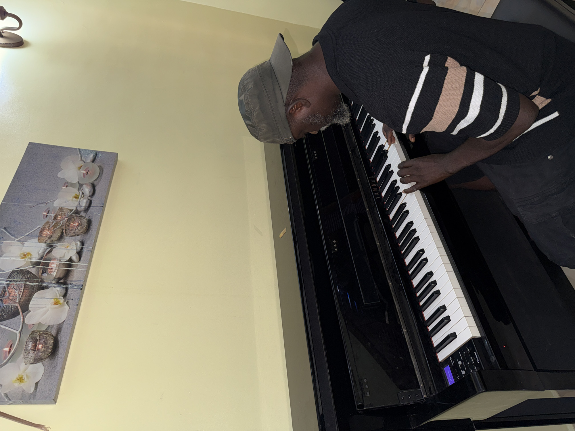 A man with gray hair and a gray cap is playing a black digital piano. He is wearing a black sweater with beige and white stripes. The piano is in front of a light yellow wall. On the wall, there is a painting with white flowers and gray stones. The location is Lekki, Nigeria.