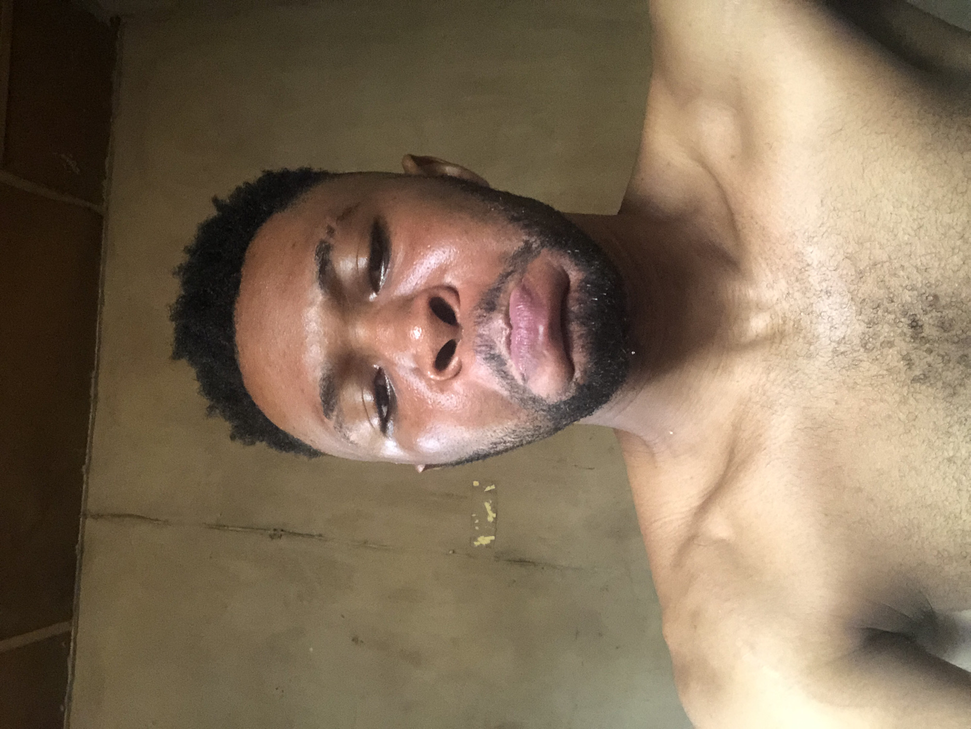 A close-up, eye-level shot shows a shirtless man with dark skin, a black beard, and close-cropped hair. His eyes are partially closed and his face appears sweaty. The background is a faded beige wall with some visible imperfections and a yellowed patch. The location is Unknown, Unknown.