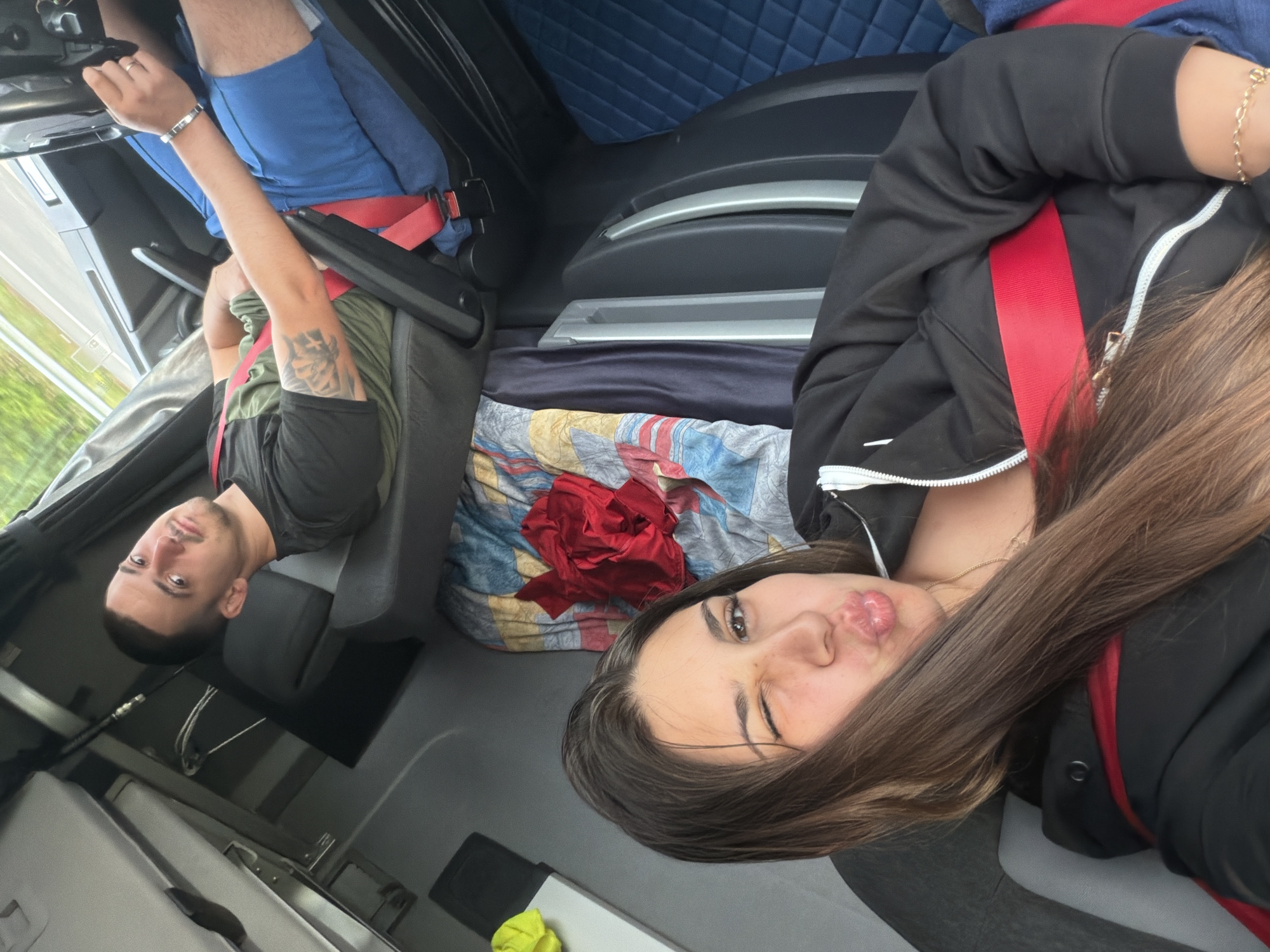Two people are inside a vehicle, likely a truck cab, as indicated by the presence of seats, steering wheel, and interior paneling. In the foreground, a woman with long brown hair is taking a selfie, puckering her lips and winking at the camera. She is wearing a black hooded jacket with a red strap across her chest and a gold necklace. Behind her, to the left, a man is seated, looking towards the camera with his head slightly tilted. He has short dark hair, a mustache, and is wearing a dark t-shirt over a green undershirt, with blue shorts. A red seatbelt is visible across his chest. Both individuals appear to be wearing seatbelts. The interior of the vehicle shows various textures and colors, including quilted blue upholstery on a seat or sleeping area, and a colorful patterned cloth spread out. The environment appears to be daytime, with some natural light filtering in, suggesting the vehicle is in motion or parked outdoors. The background shows a blurred, out-of-focus view of greenery and a road, further supporting the idea that they are in a vehicle.