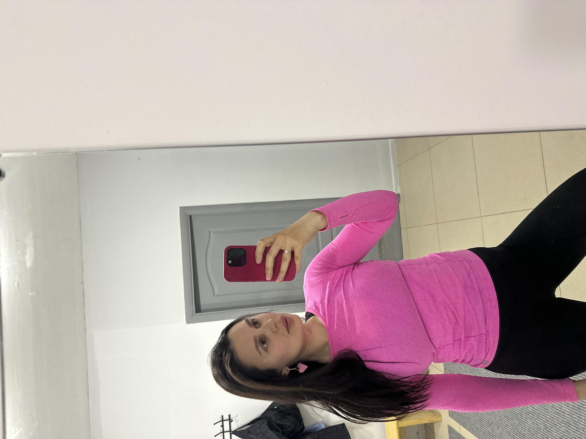 A woman takes a mirror selfie indoors. She is wearing a bright pink long-sleeved top and black leggings. She holds a red cell phone in her right hand. She has long dark hair and wears pink earrings. The reflection shows a grey door and a white wall. The floor has beige tiles. A clothes rack is visible in the lower left corner. The location is Chaikovsky, Russia.