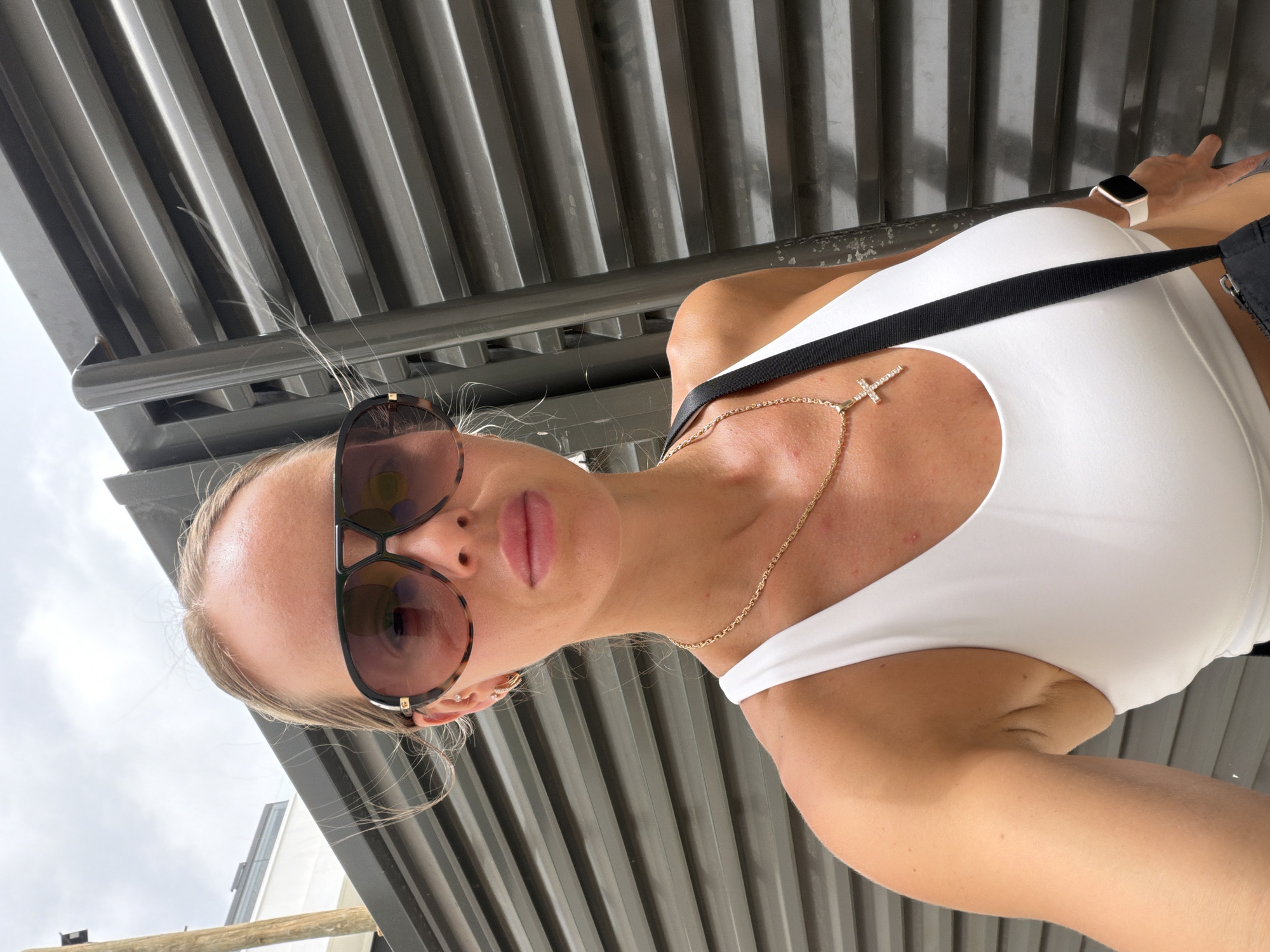A close-up selfie of a woman with blonde hair pulled back is taken outdoors. She is wearing large, tortoise-shell aviator sunglasses, a white tank top, and a gold chain necklace with a cross pendant. A black strap from a bag is visible across her shoulder, and a white smartwatch is on her left wrist. She is standing in front of a dark grey, corrugated metal fence. The background suggests an urban environment, possibly in Finestrat, Spain, with a hint of a building to the left. The lighting is overcast, indicating a cloudy day.