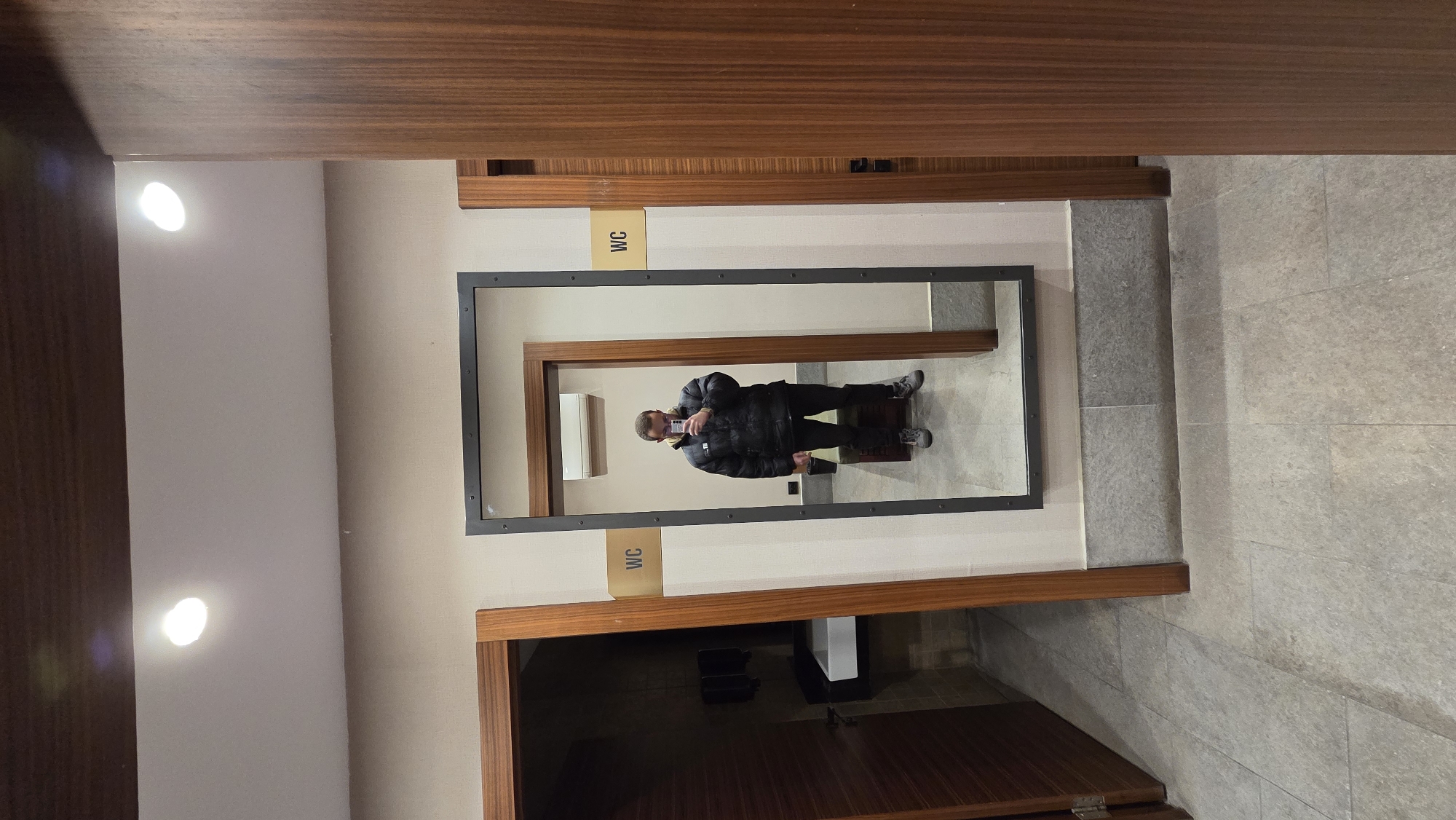 A full body reflection of a man taking a selfie is visible in a mirror in a public restroom. The man is wearing a black puffy coat and dark pants, with gray shoes. He is holding a smartphone up to take the picture. Behind him is a light-colored wall and floor, with some dark fixtures. The mirror is framed in gray and is set within a wooden doorway. There is a sign saying "WC" above the doorway. The setting appears to be a restroom in Bania, Bulgaria.