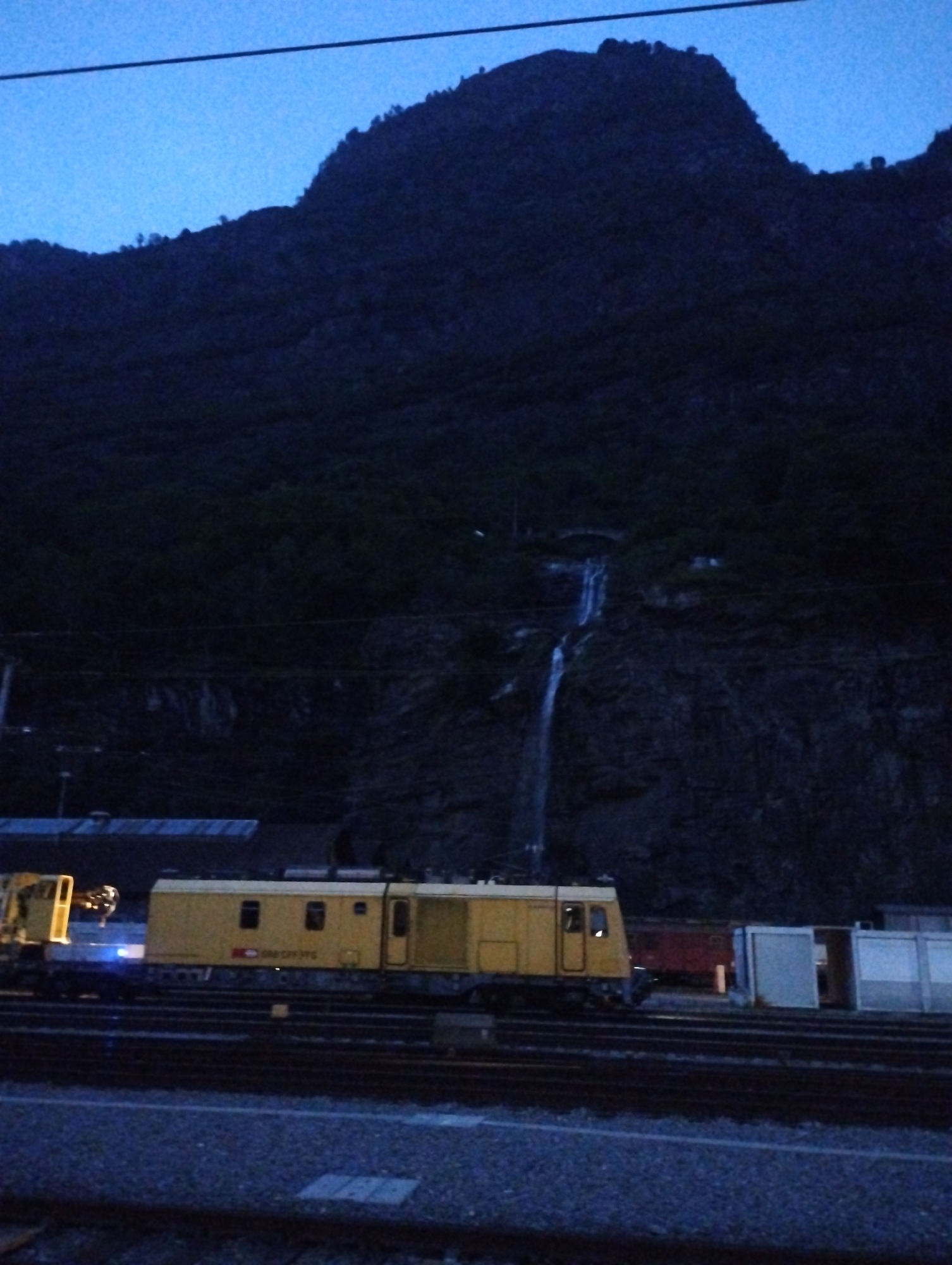 The image depicts a mountainous landscape at dusk, with a dark, silhouetted mountain range dominating the background. A thin waterfall cascades down the rocky face of the mountain. In the foreground, a yellow train engine is parked on railway tracks. The train appears to be a work train, possibly for maintenance or construction, indicated by its utilitarian design and the presence of what looks like equipment attached to it. Several other railcars and structures are visible further down the tracks, also partially obscured by the dim lighting. The sky is a deep twilight blue, suggesting the time is either early evening or pre-dawn. The overall scene conveys a sense of solitude and industrial activity set against a natural backdrop.