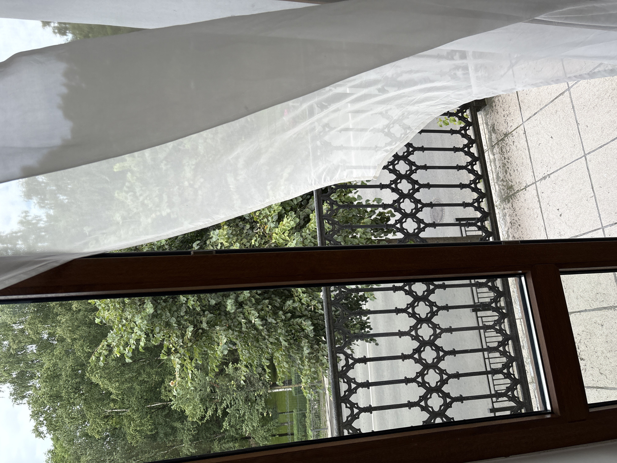 An eye-level, medium outdoor shot captures a view from a balcony looking out towards a tree-lined street in Gatchina, Russia. In the foreground, the edge of a wooden door frame on the left and a white sheer curtain billowing on the right frame the scene. Beyond the balcony railing, a grey asphalt street is visible with a black ornamental metal fence separating it from a green grassy area and more trees. The overcast sky suggests a cloudy day.