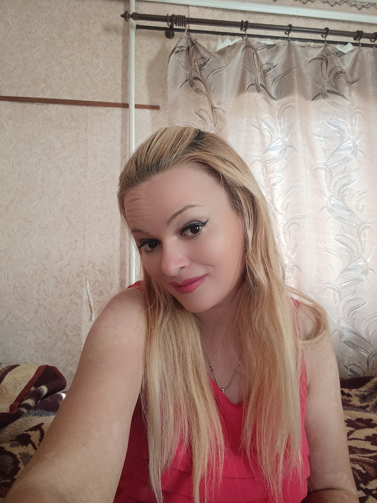 A blonde woman with fair skin and bright pink lipstick is pictured in a selfie. She has on a pink top and a silver necklace. Her hair is long and reaches her shoulders. The background appears to be an indoor setting with patterned wallpaper and curtains. There are also hints of bedding or upholstery visible, suggesting a bedroom or living area. No specific landmarks or indicators for Chernihiv, Ukraine are present in the image.