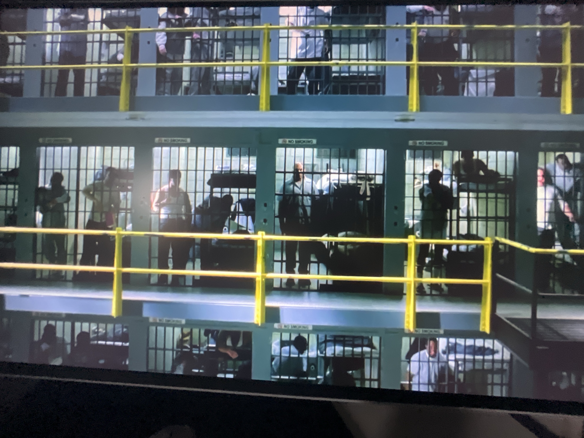 The image shows a prison block with multiple cells visible on different levels. The cells have metal bars and some have beds inside. Several individuals, presumably inmates, are visible within the cells, some standing and some lying down. The environment appears to be an indoor correctional facility. There is no discernible information within the image that would indicate its location in the city of Varna, Bulgaria.
