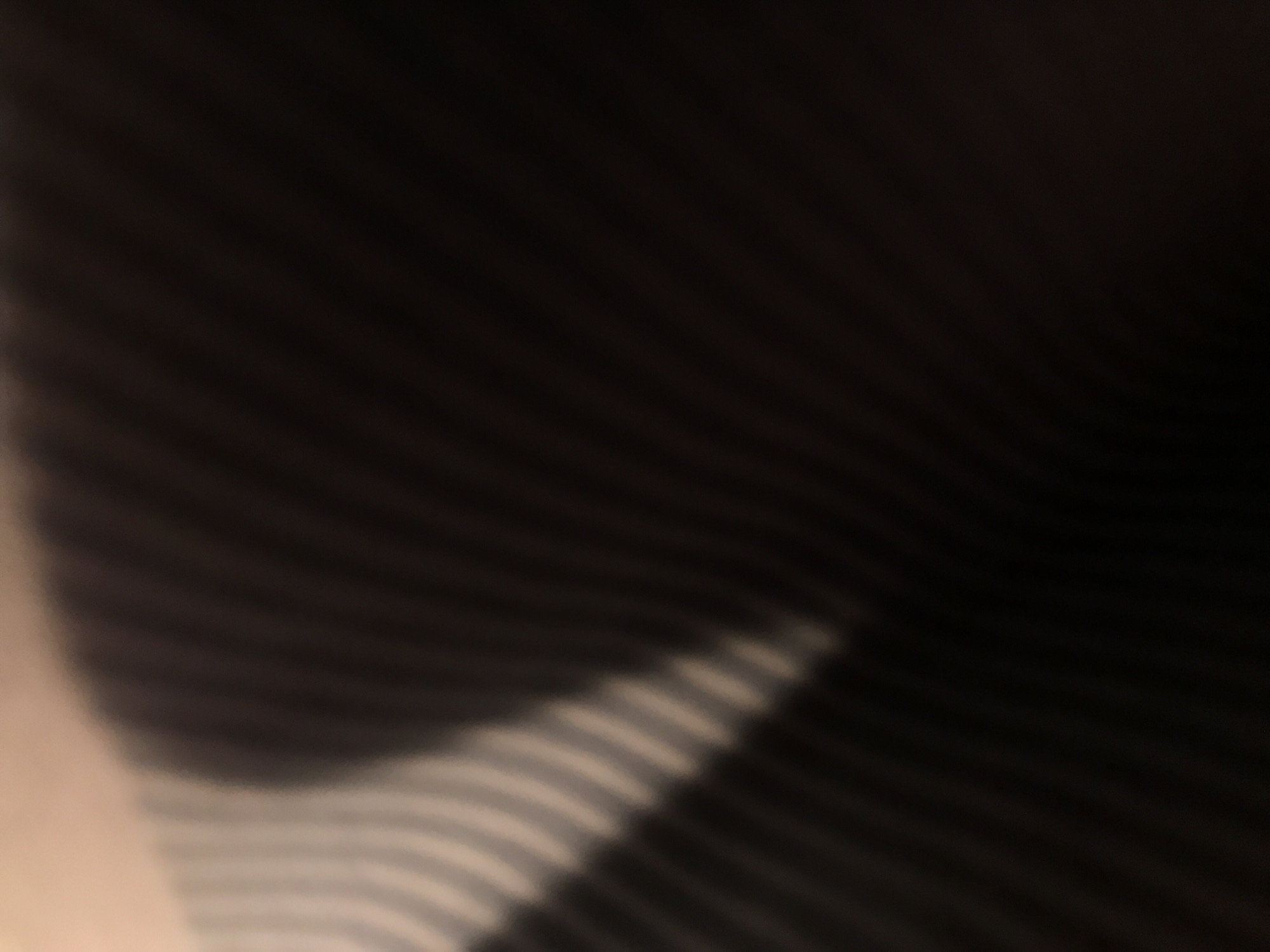 The image shows a blurry, close-up view of a textured surface. The surface appears to consist of parallel lines or ridges, alternating between light and dark shades. The lighting is uneven, with the lower left corner brighter and the upper right corner darker. The image is out of focus, making it difficult to discern the exact material or texture. There is no information to suggest this image was taken in Yerevan, Armenia.