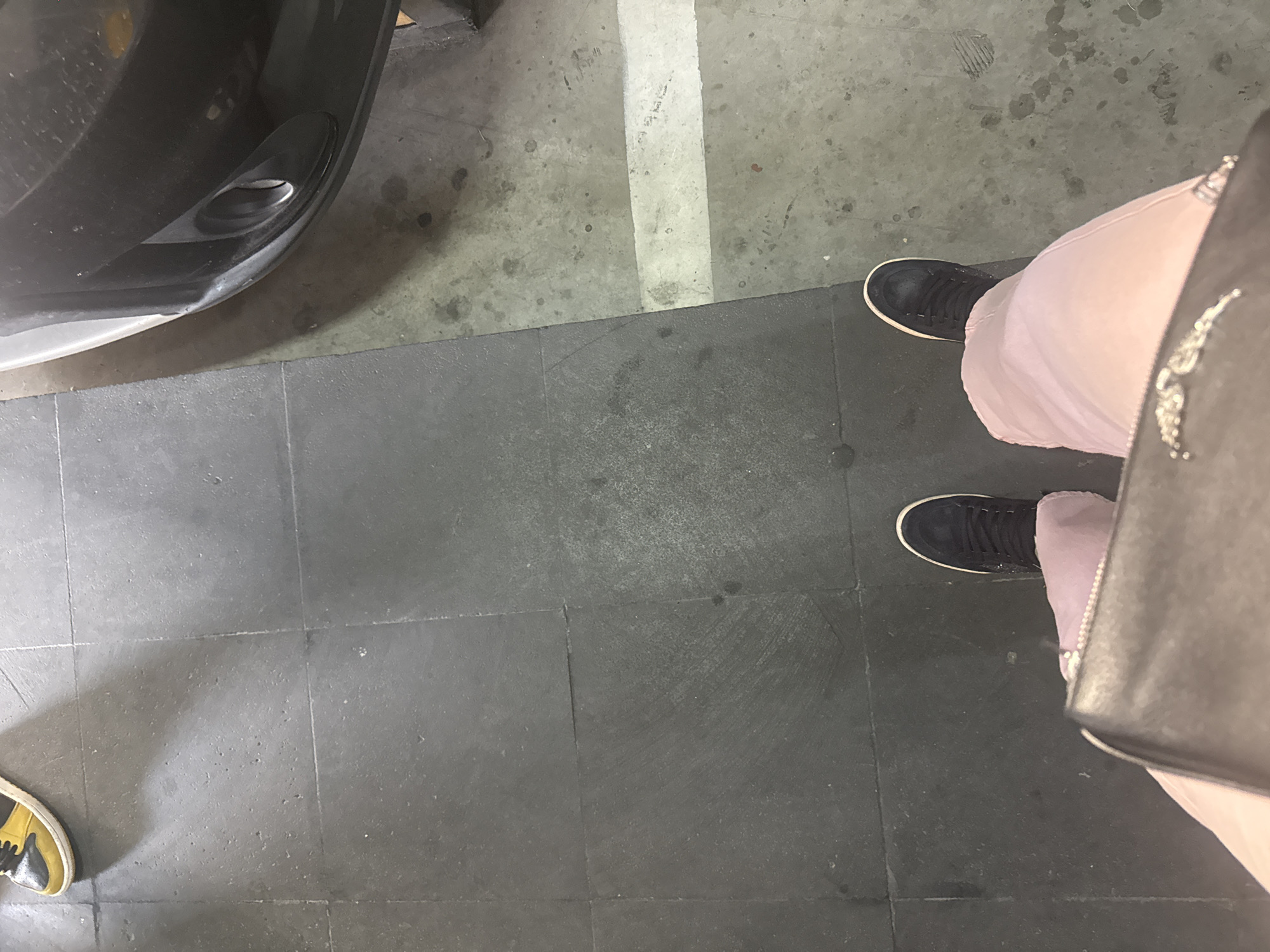 An overhead shot captures a ground-level view in what appears to be an indoor parking area in San Bernardino Tlaxcalancingo, Mexico. The scene is dominated by two distinct floor surfaces: rough, grey concrete with a faded white parking line, and smooth, dark grey square tiles.
On the right side of the image, two feet of a person are visible, wearing black sneakers with white soles. This person is wearing light pink or purple trousers, and a black bag or purse with a textured pattern (possibly studded or embroidered) is partially visible hanging next to their right leg. The left foot is slightly further away, while the right foot is closer to the bottom right corner.
In the upper left corner, the front bumper of a dark-colored car (possibly black or dark grey) is visible, with a circular element that might be a fog light or sensor. The car is positioned on the concrete section of the ground.
Another foot, wearing a yellow and black sneaker with a white sole, is partially visible in the bottom left corner, resting on the dark grey tiled floor.
The transition between the concrete and the dark grey tiles creates a sharp, horizontal dividing line across the middle of the frame. The concrete section shows various stains and small specks, along with faint, illegible scribbling or scuff marks on and around the white line. The tiled area also shows some minor scuffing and imperfections. The lighting is somewhat dim, suggesting an indoor or covered environment, and creates soft shadows. There is no clear activity, with the individuals appearing to be stationary or mid-stride.